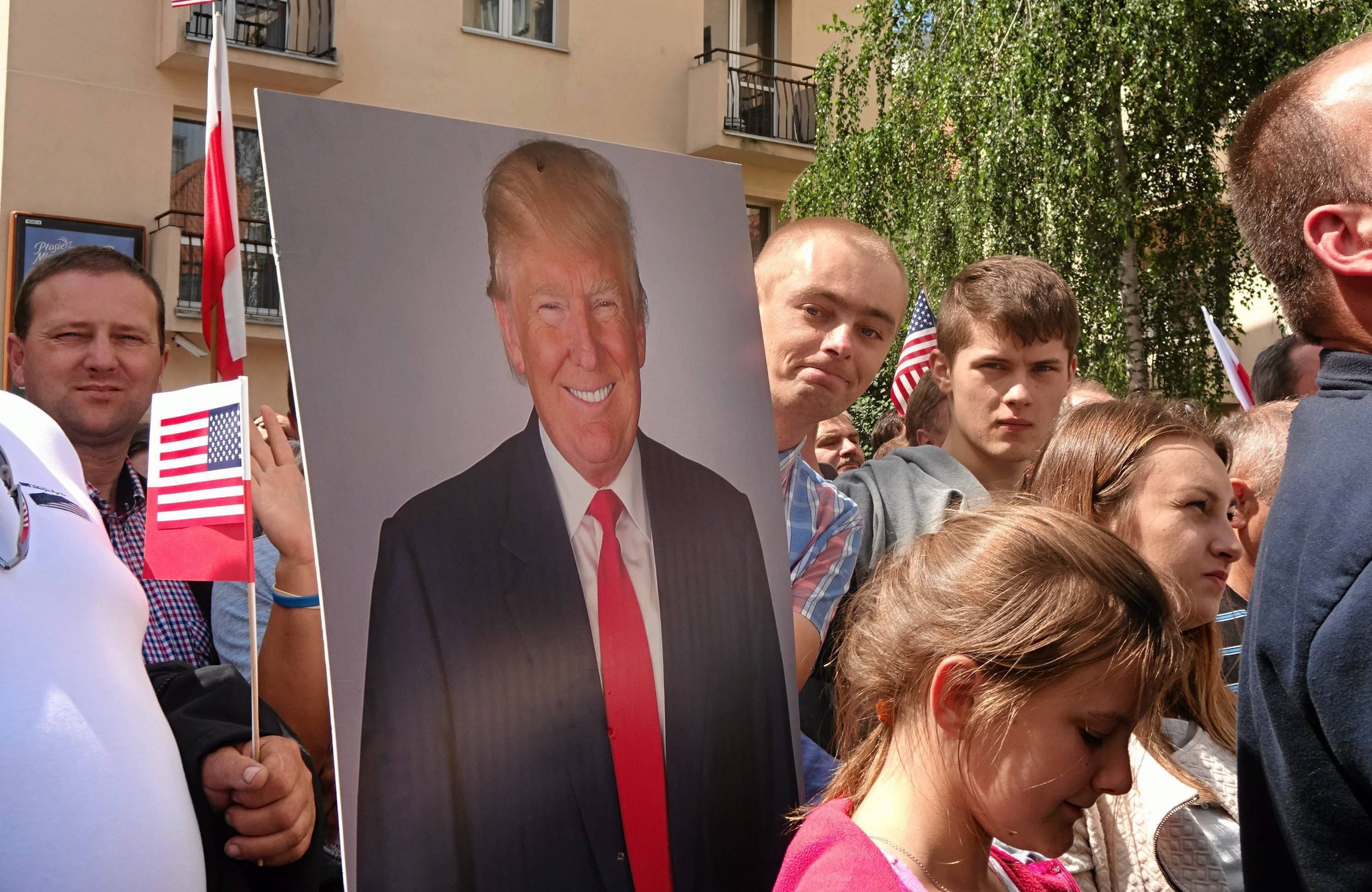 Na zdjęciu widać tłum ludzi stojący na ulicy. Jedna osoba trzyma małe flagi - polską i amerykańską, inna duży portret (obraz) Donalda Trumpa