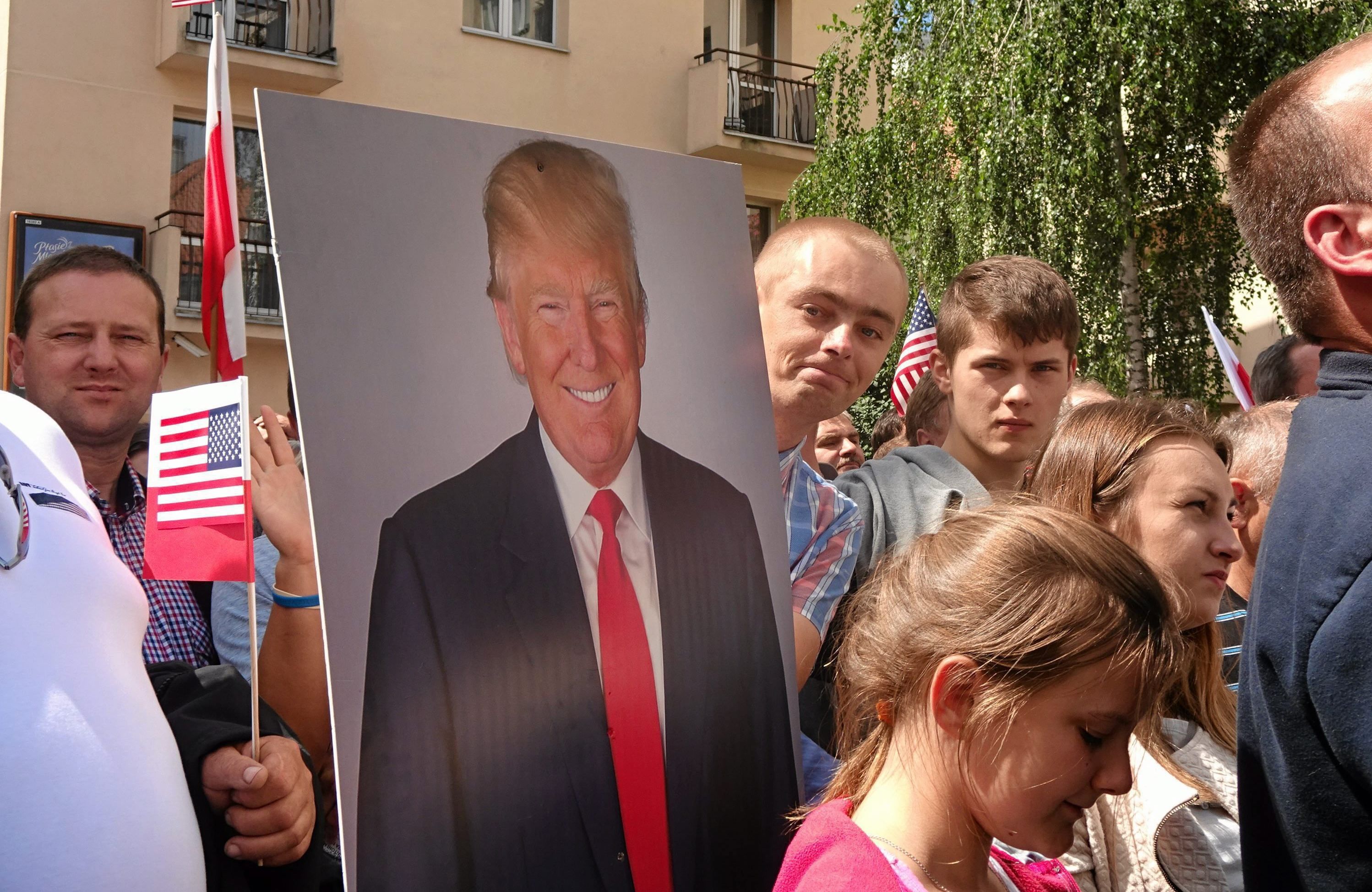 Na zdjęciu widać tłum ludzi stojący na ulicy. Jedna osoba trzyma małe flagi - polską i amerykańską, inna duży portret (obraz) Donalda Trumpa