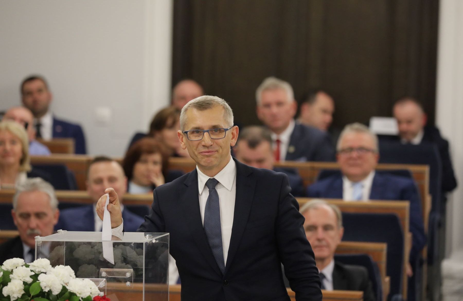 siwiejący mężczyzna w garniturze i okularach na tle ław senackich