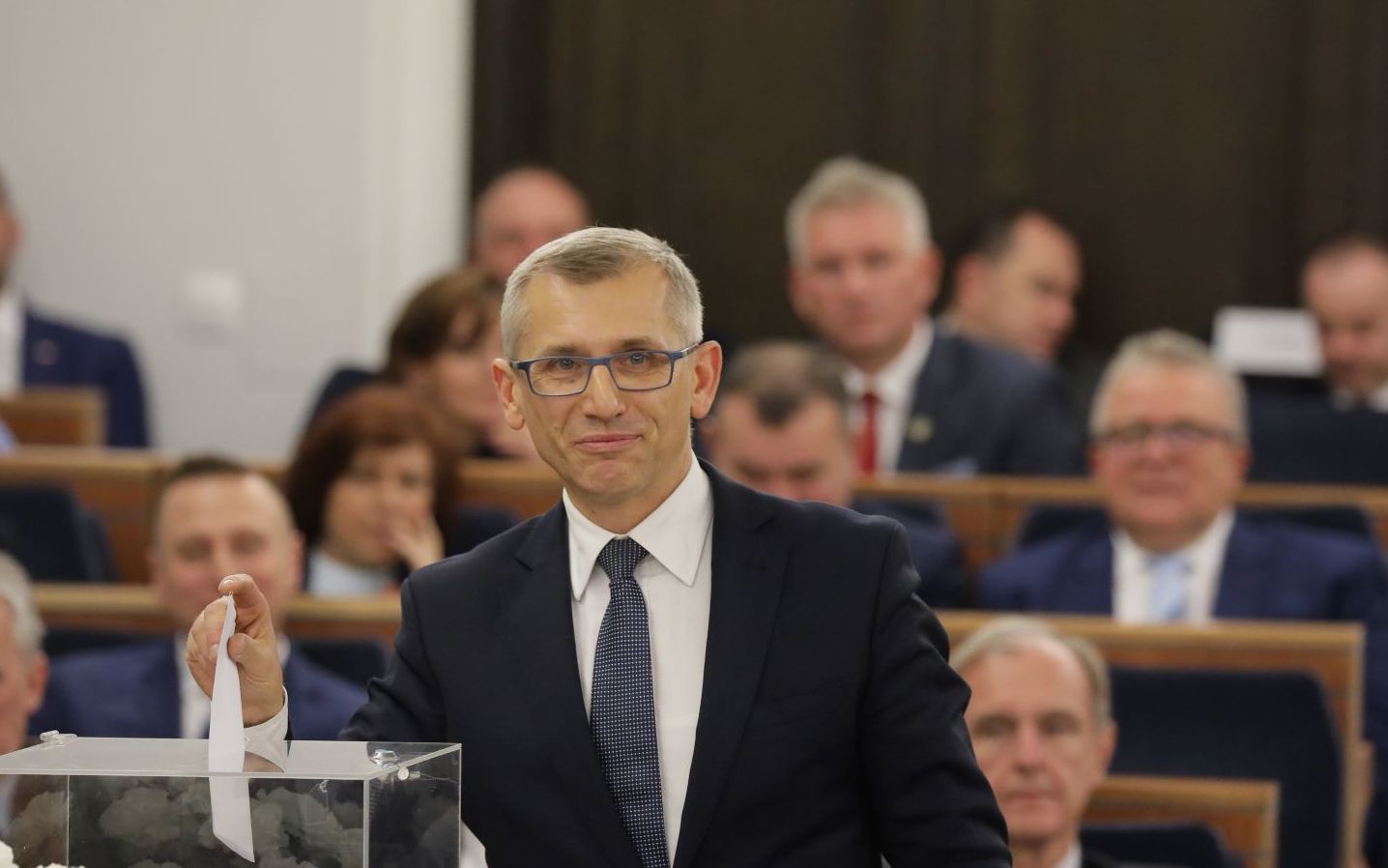 siwiejący mężczyzna w garniturze i okularach na tle ław senackich