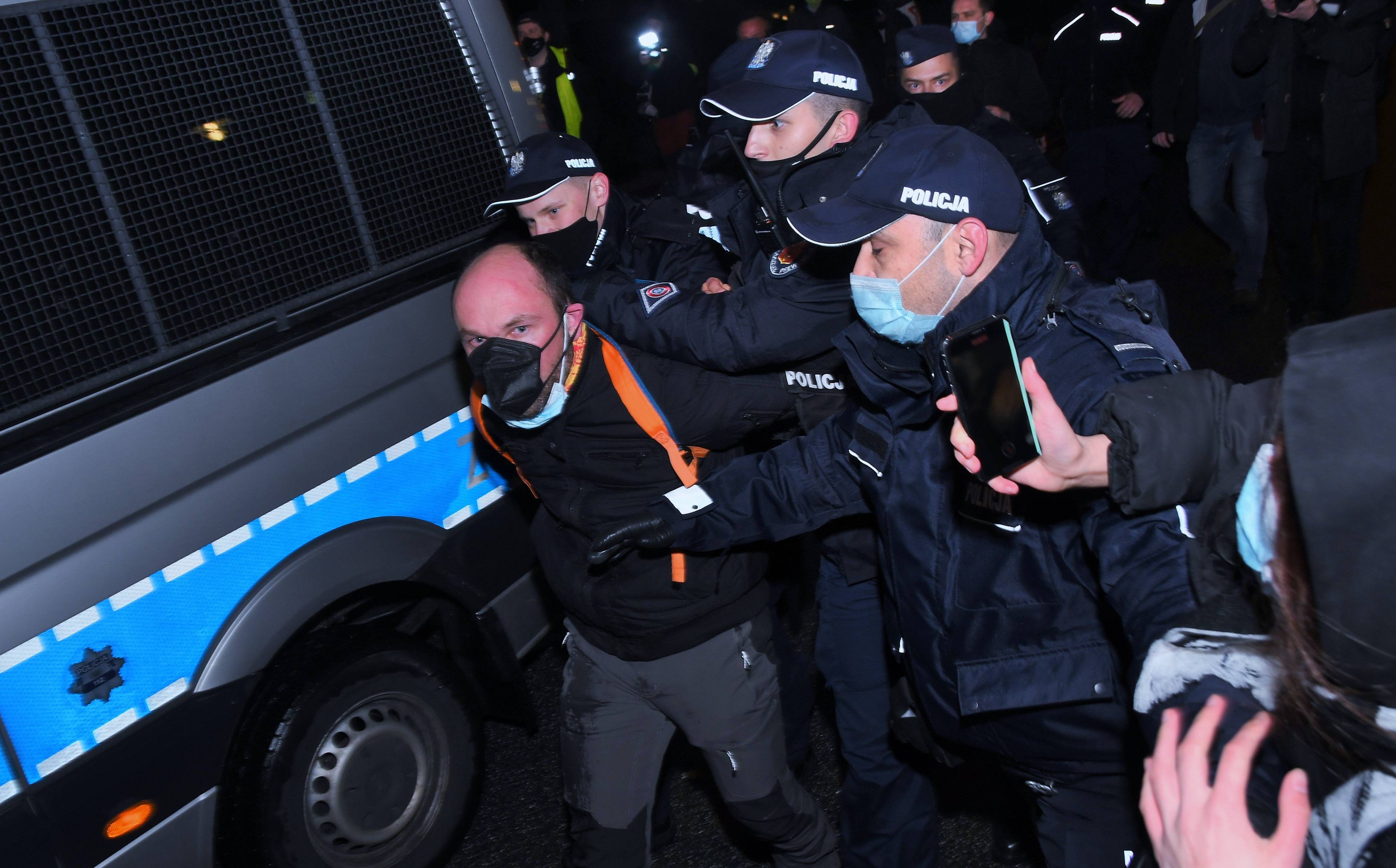 Na zdjęciu aktywista Dominik Berliński. Mężczyzna w maseczce, zatrzymany przez umundurowanych policjantów. Policjanci prowadzą go do radiowozu. Mężczyzna ma ręce skute z tyłu.