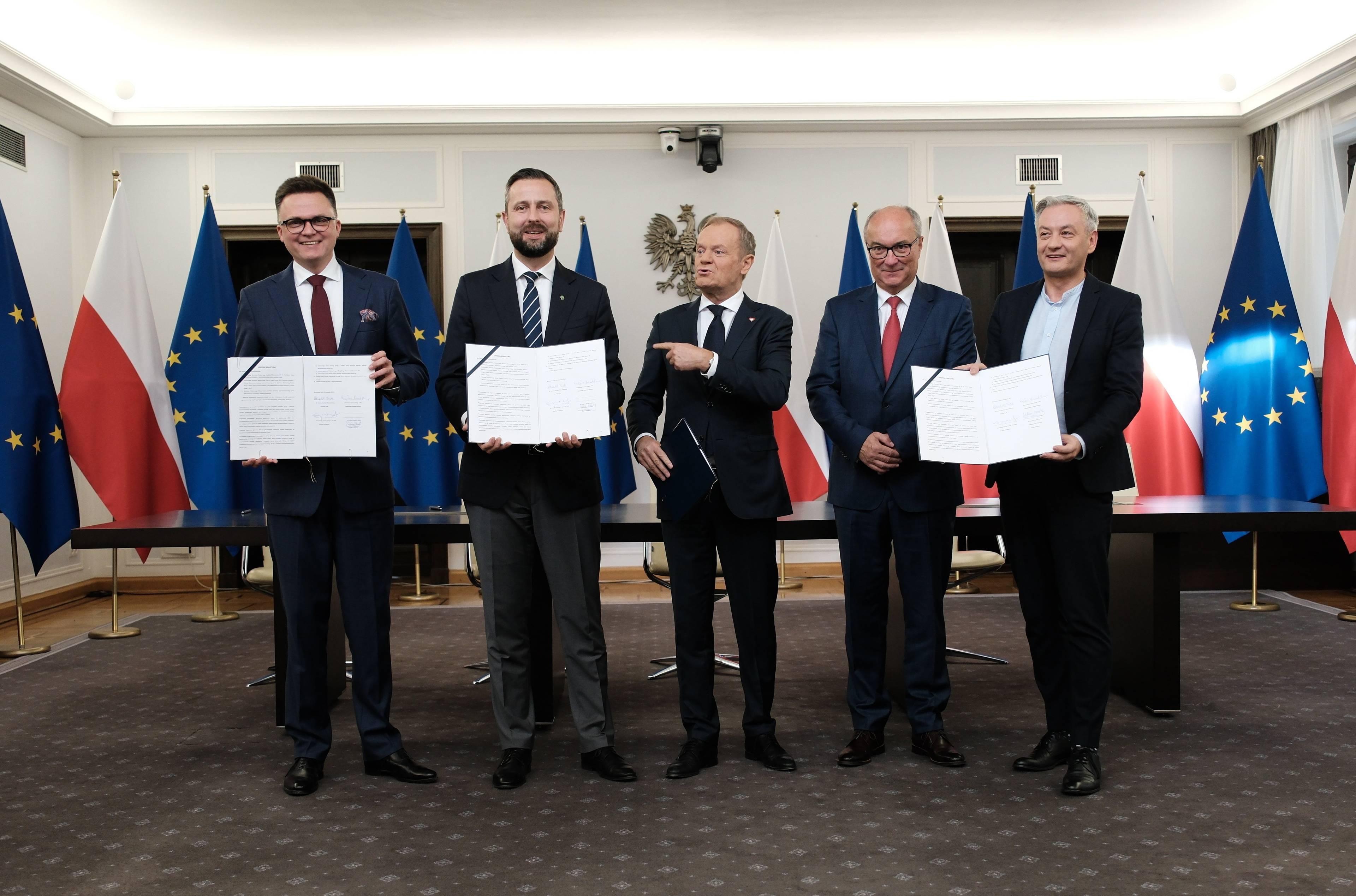 Liderzy Trzeciej Drogi, KO i Nowej Lewicy stoją obok siebie, trzymając podpisaną umowę koalicyjną, w tle flagi Polski i Unii Europejskiej
