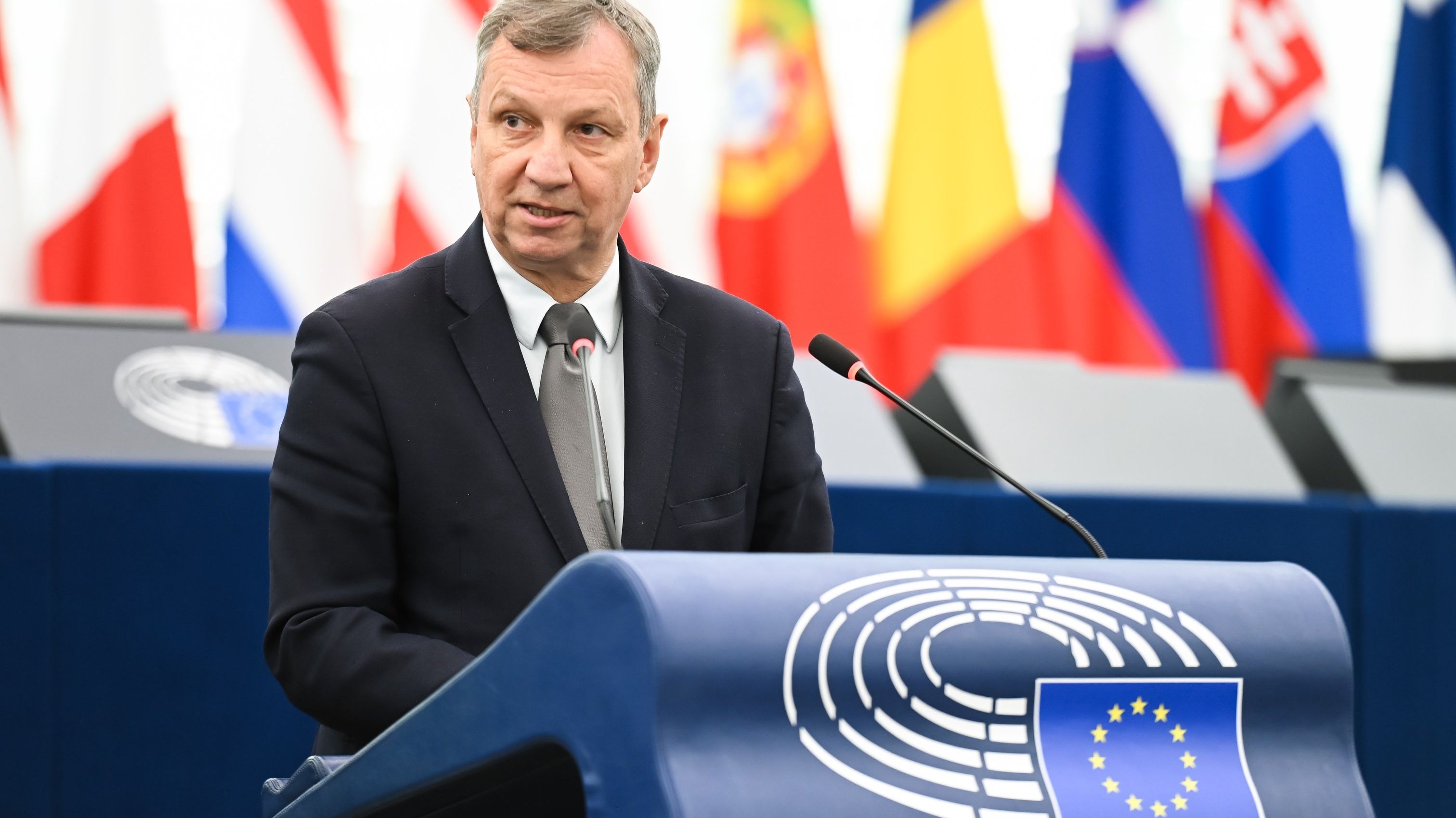 Mężczyzna w garniturze przemawia z mównicy Parlamentu Europejskiego. Za nim widać flagi państw UE i logo Unii Europejskiej. Na sali obecni są inni uczestnicy obrad.