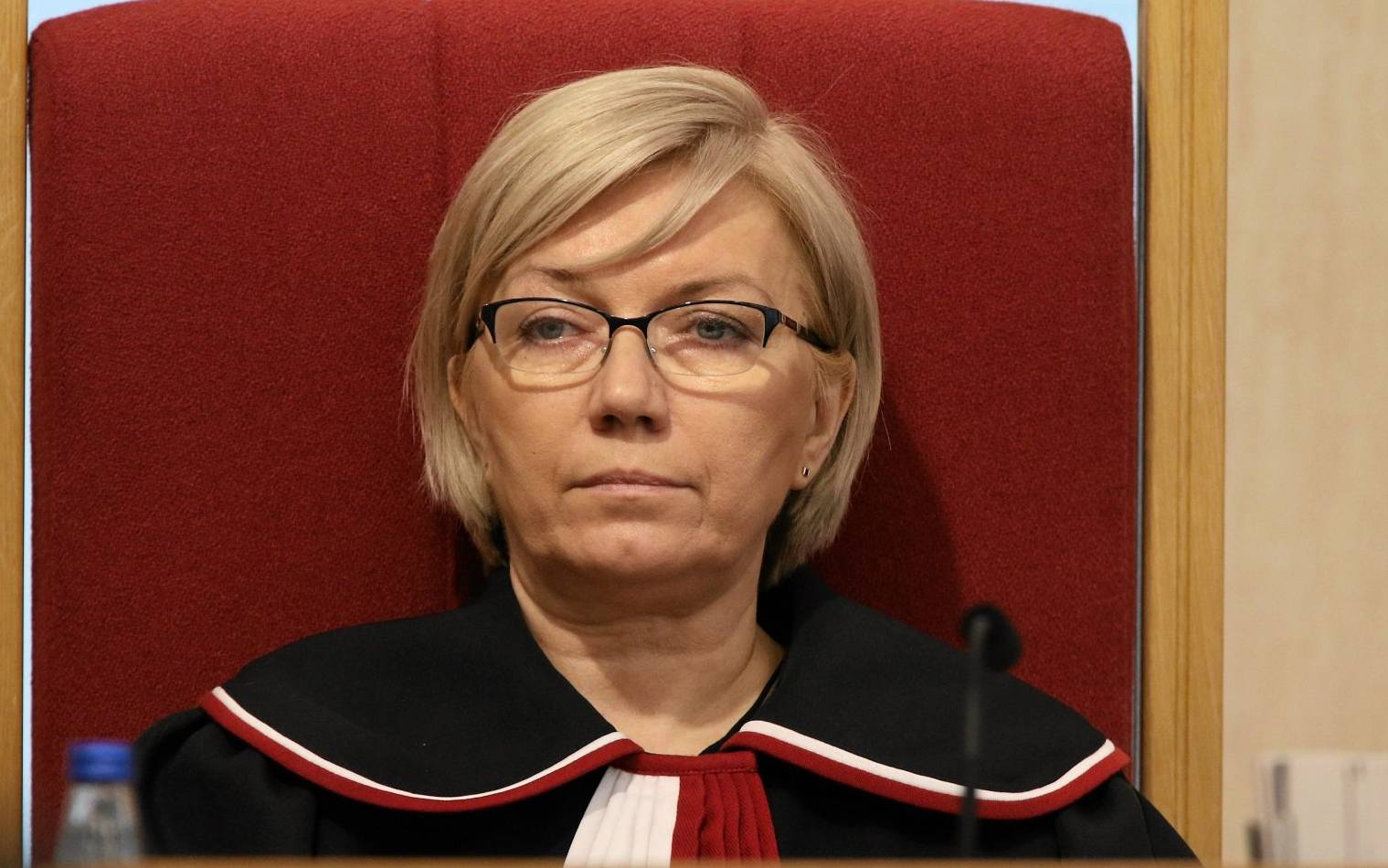 Julia Przyłębska, blondynka z włosami średniej długości, w okularach i w todze sędziowskiej siedzi w czerwonym fotelu
