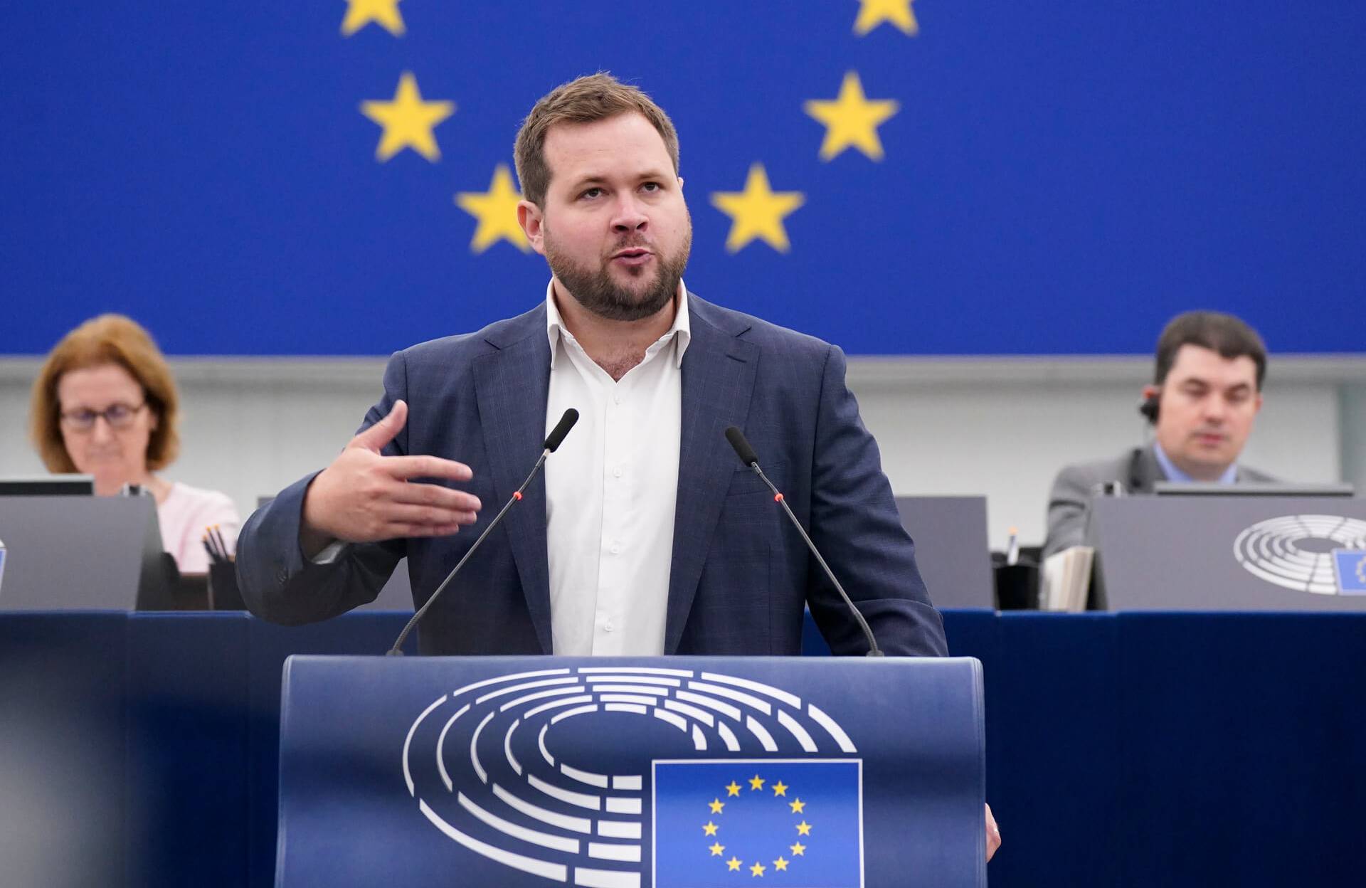 Mężczyzna z krótkimi włosami ciemny blond i krótką brodą stoi za mównicą w sali plenarnej Parlamentu Europejskiego, w tle za nim europejska flaga z gwiazdkami, gestykuluje prawą ręką i przemawia do zebranych europosłów.