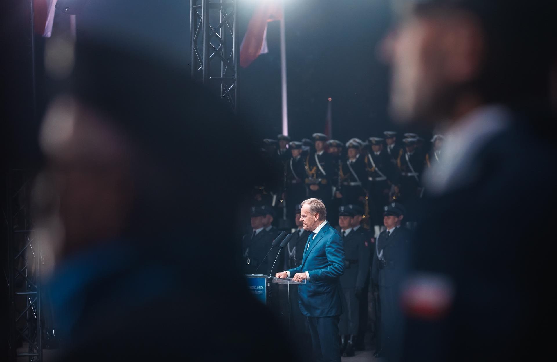Mężczyzna w niebieskim garniturze stoi przy mównicy z mikrofonem, widziany z daleka, stoi na tle wojskowych w mundurach marynarki wojennej, jest ciemno, świecą się sztuczne światła.