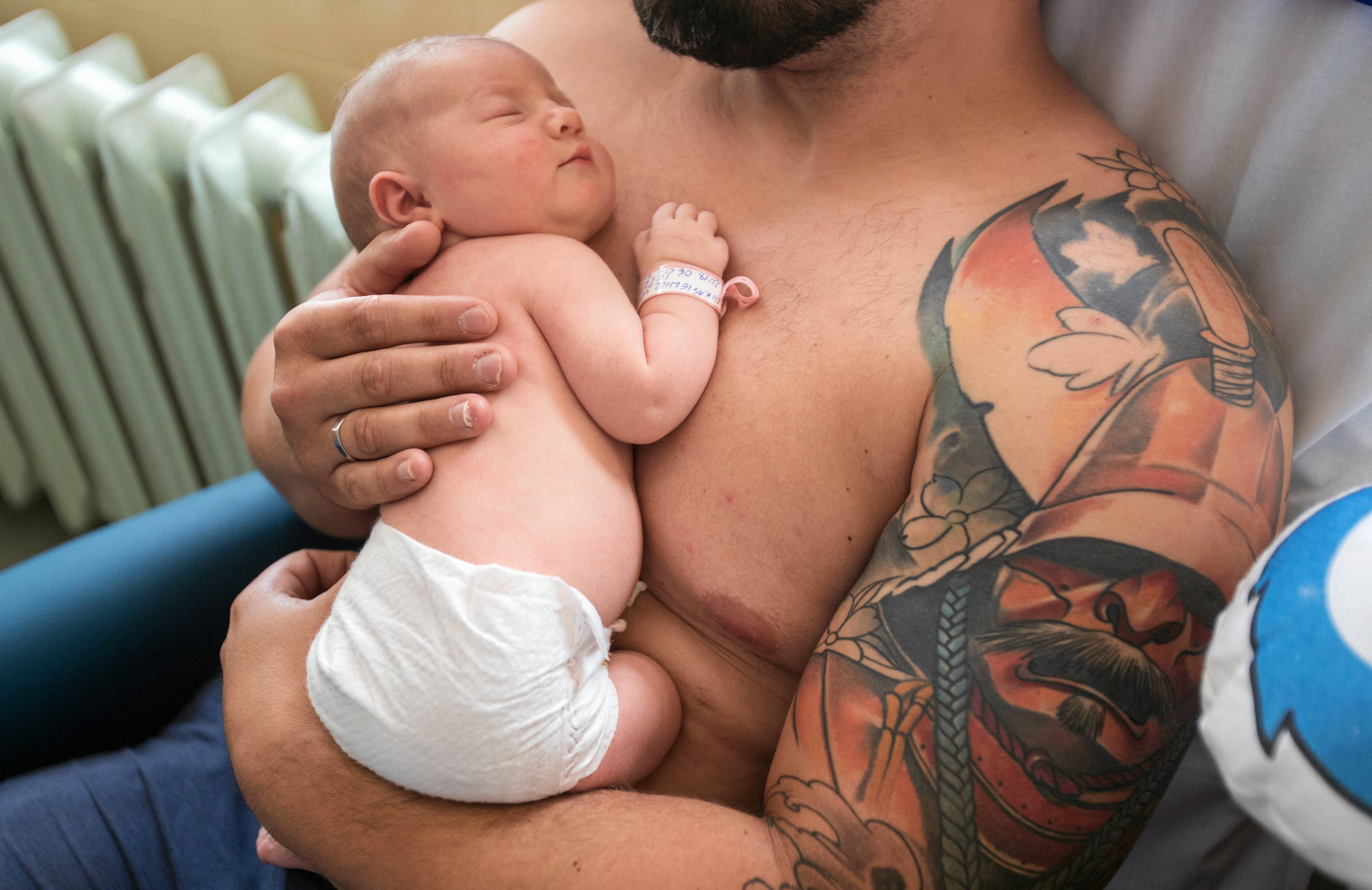 Mężczyzna z nagim torsem przytula noworodka. Na lewym ramieniu ma tatuaż przedstawiający wojownika