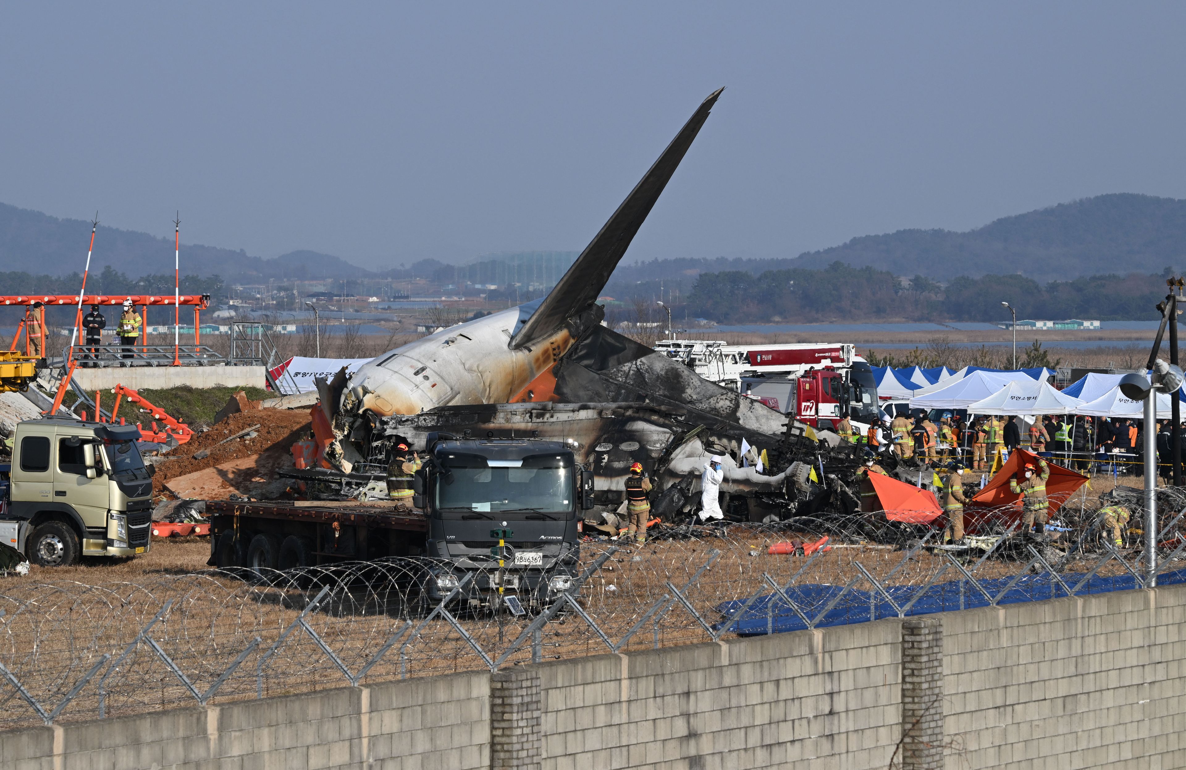 Zdjęcie katastrofy samolotu Jeju Air na lotnisku w Muan w Korei Południowej. Widzimy zniszczoną, spaloną maszynę leżącą na pasie startowym, tylko ogon zachował jeszcze swój pierwotny kształt. Wokół samolotu są służby porządkowe
