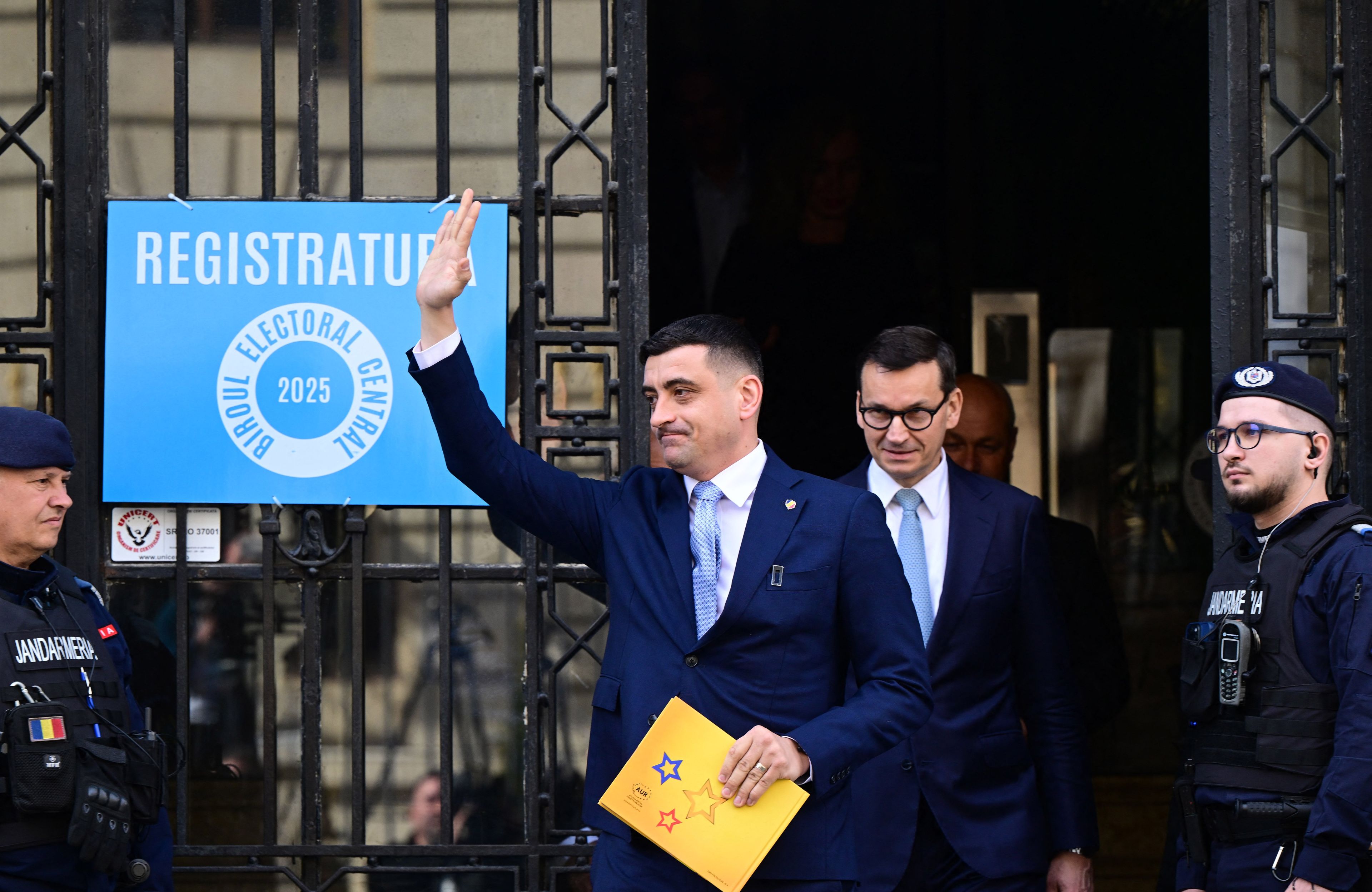 Dwóch mężczyzn wychodzi z siedziby komisji wyborczej, ten z porzudu podnosi rękę , ten z tyłu, Morawiecki, uśmiecha się