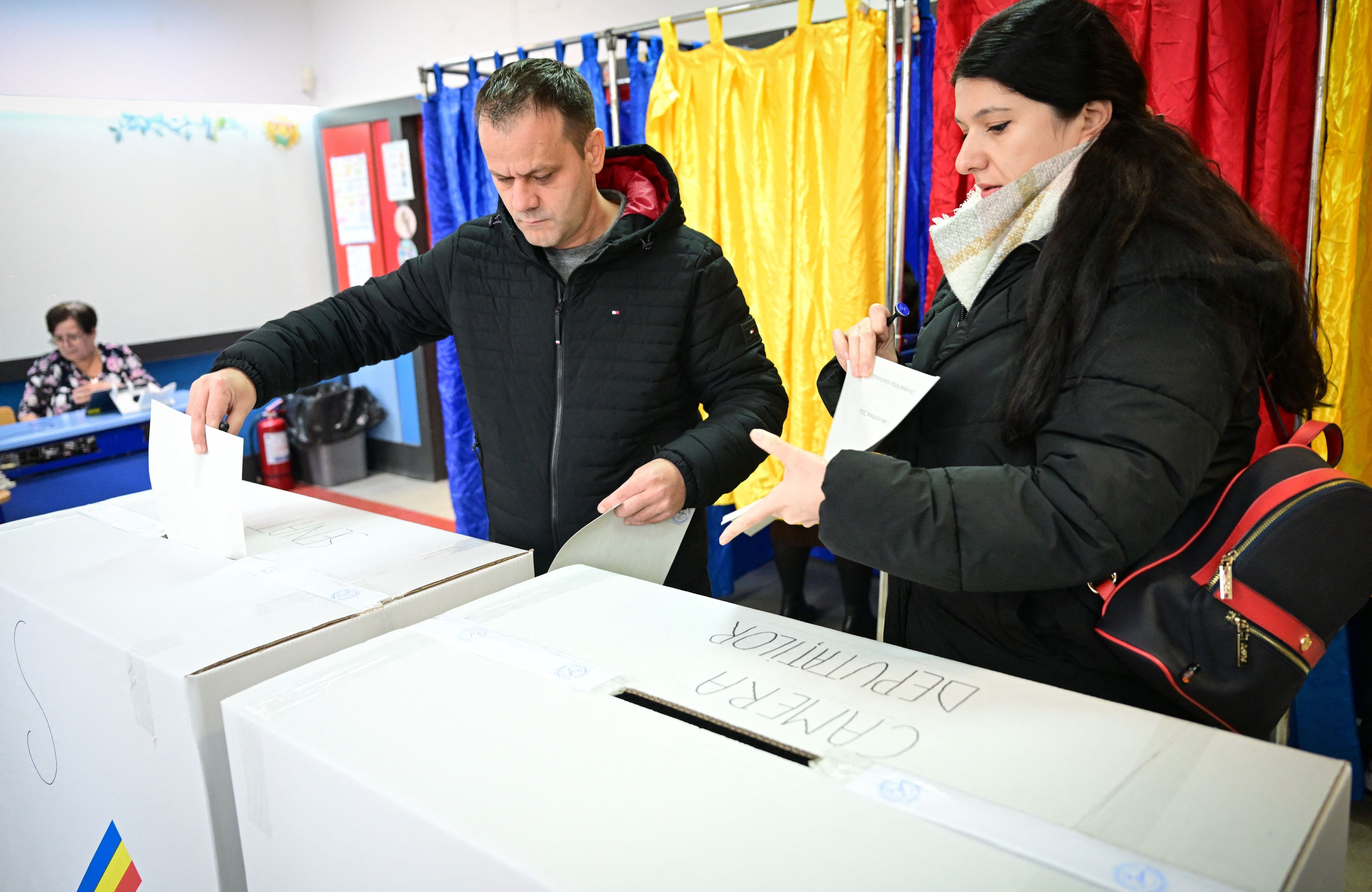 Ludzie wrzucają głosy do urn. W tle - pomieszczenia do zakreślania głosów z kotarami w kolorach rumuńskiej flagi: niebieskim, żółtym i czerwonym