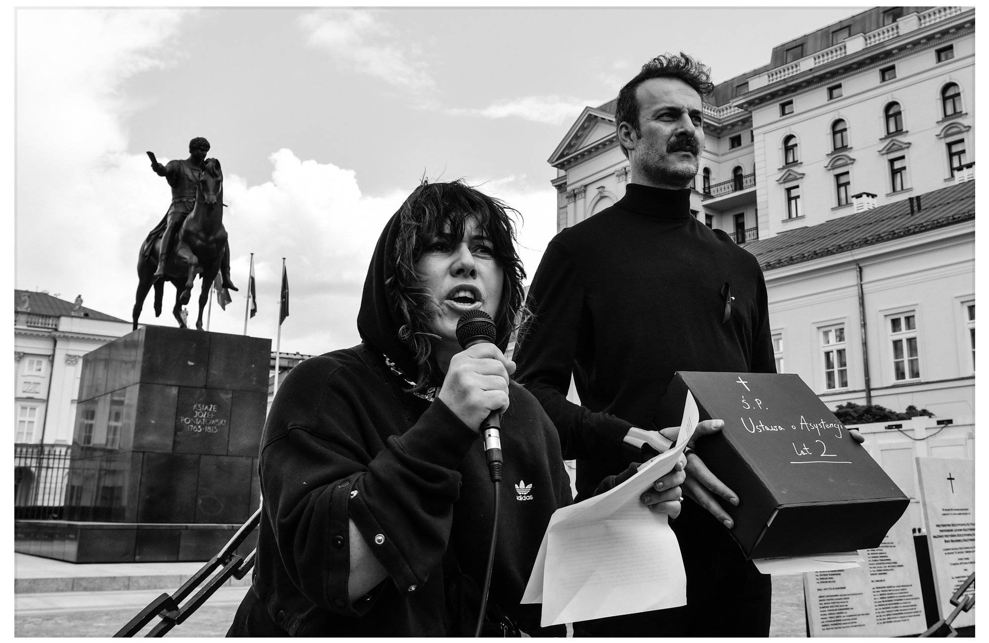 Czarno-białe zdjęcie: kobieta (Agnieszka Szlipa) przemawia pod Pałacem Prezydenckim w sprawie osób z niepełnosprawnościami