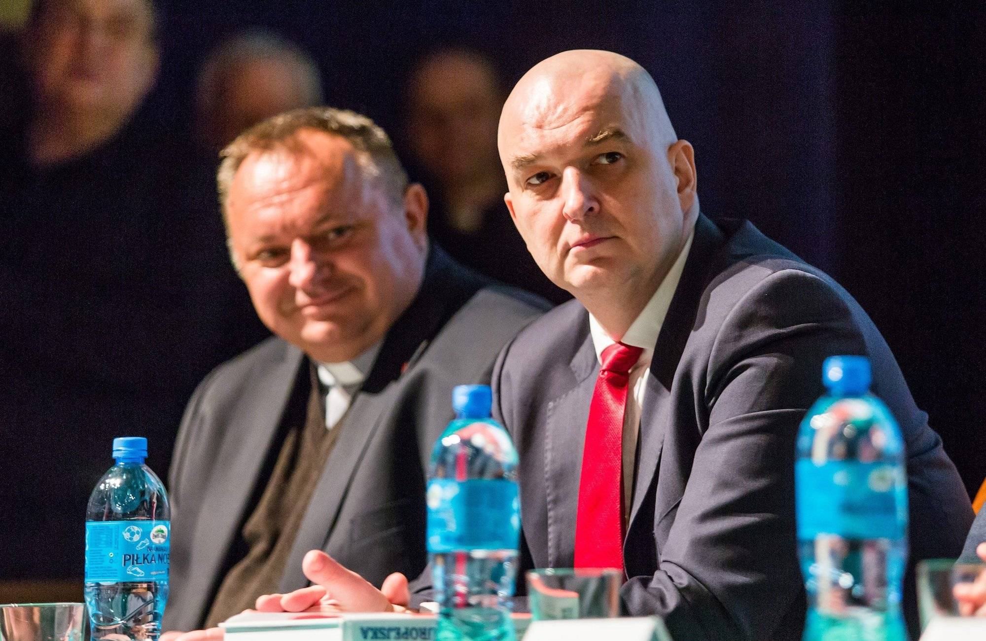 Sławomir Dębski w ciemnym garniturze i czerwonym krawacie siedzi przy stole podczas konferencji. Obok niego ks. Waldemar Cisło.
