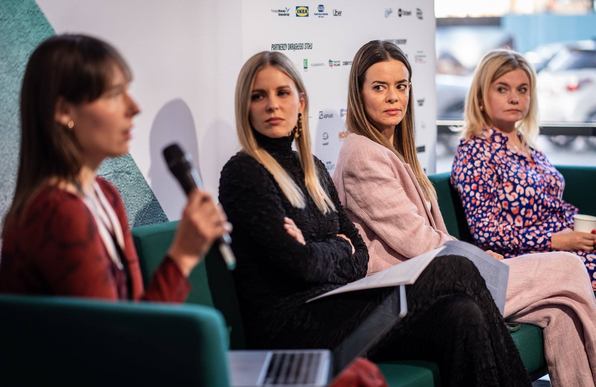 Cztery kobiety siedzą w panelu, pierwsza z lewej trzyma mikrofon, pozostałe patrzą na nią z poważnymi minami