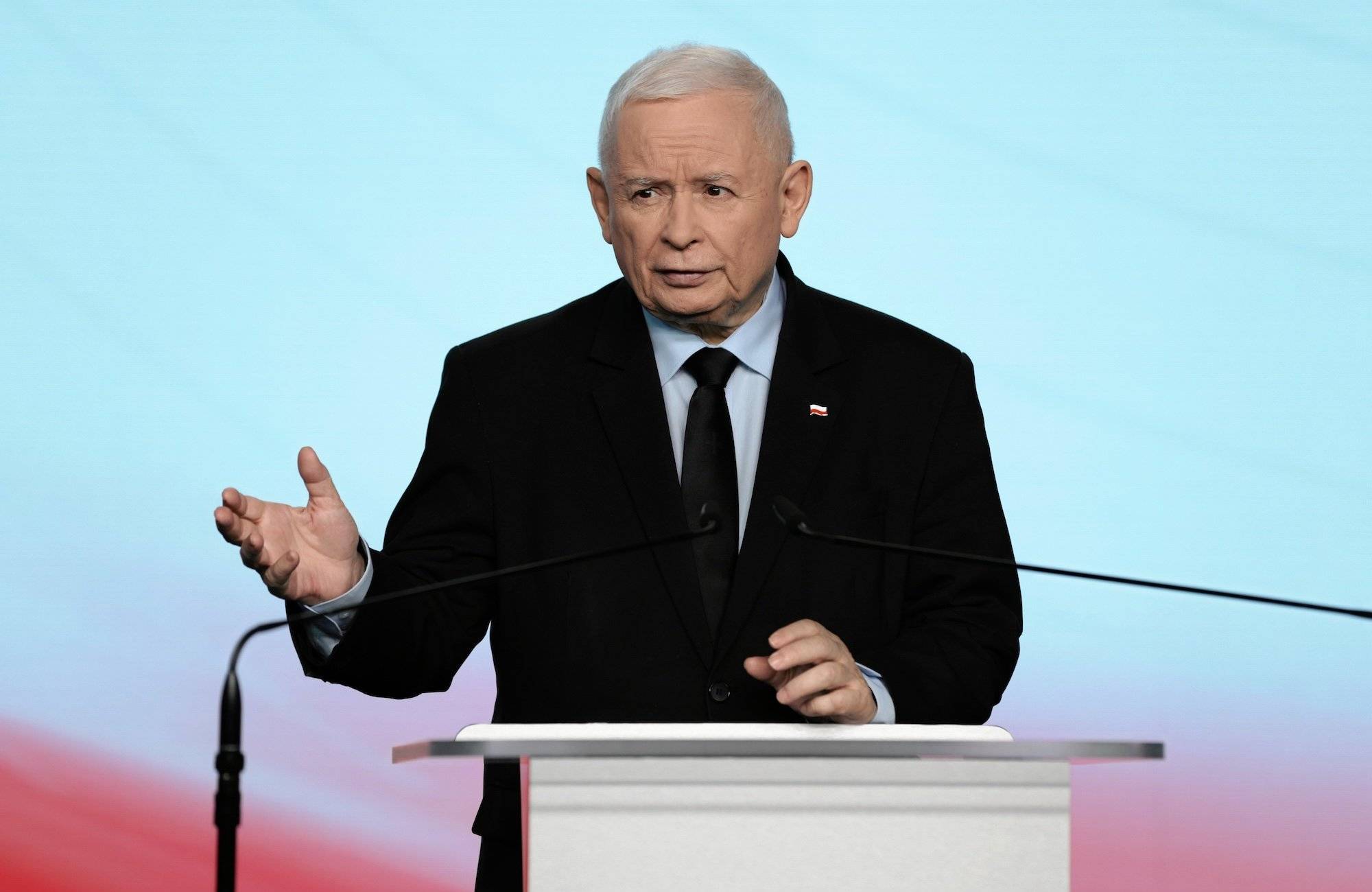 Jarosław Kaczyński podczas wystąpienia. Przemawia zza pulpitu i unosi prawą rękę.