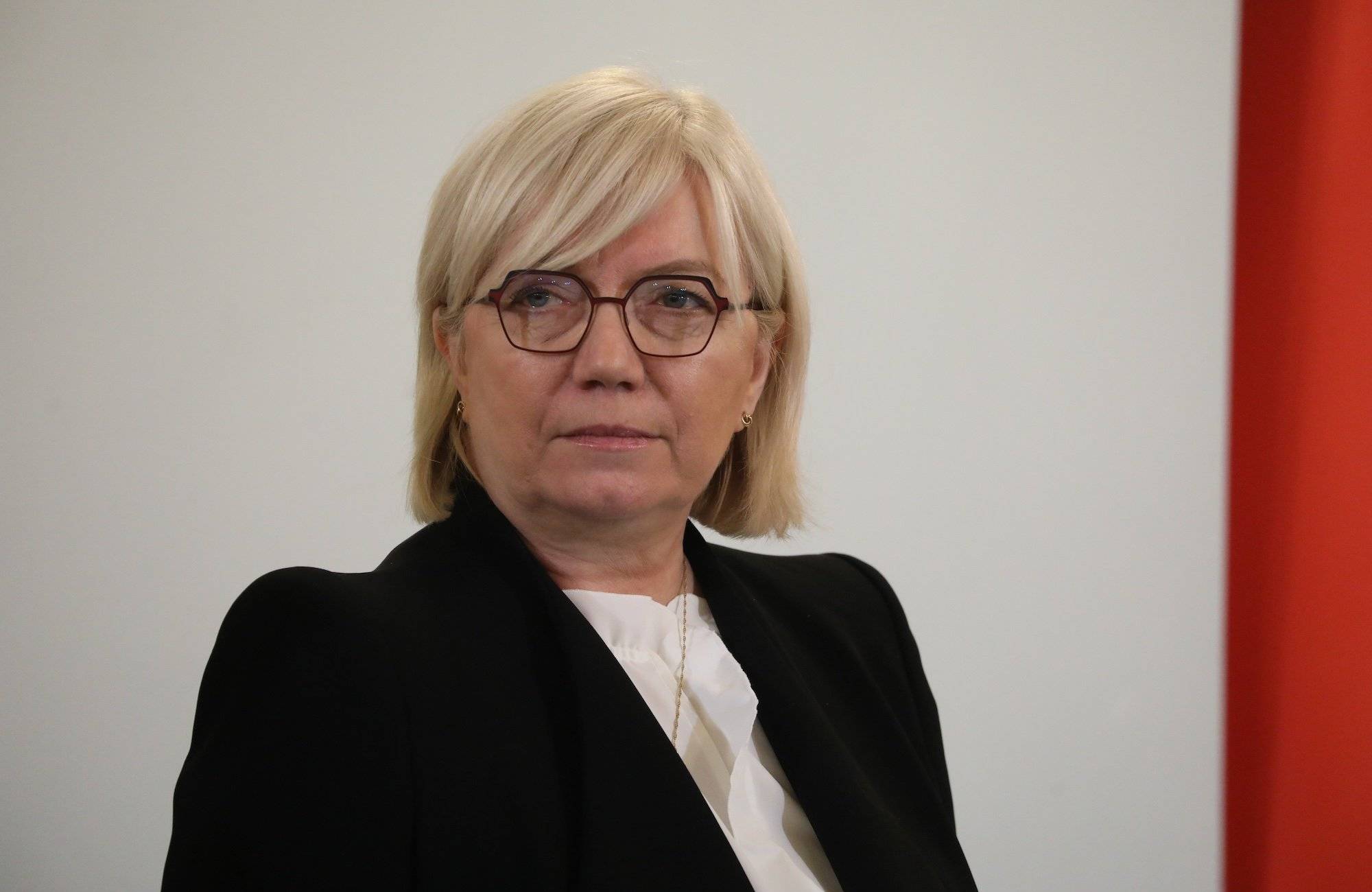 Portret Julii Przyłębskiej, ubranej w białą bluzkę i czarną marynarkę. Ma okulary w ciemnych oprawkach i jasne włosy