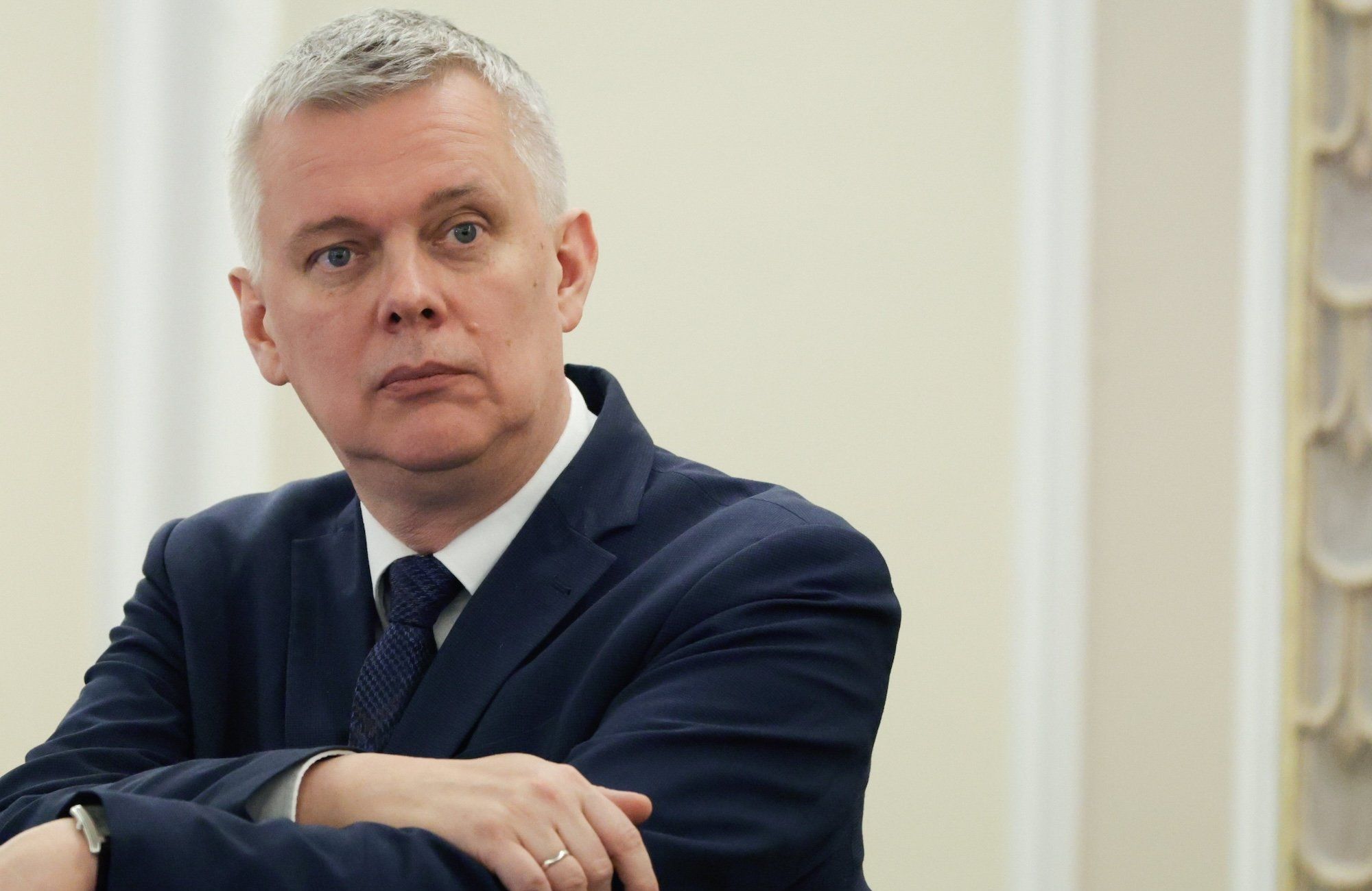 Tomasz Siemoniak w ciemnym garniturze patrzy w dal z zaniepokojoną miną, upierając się rękami o krzesło