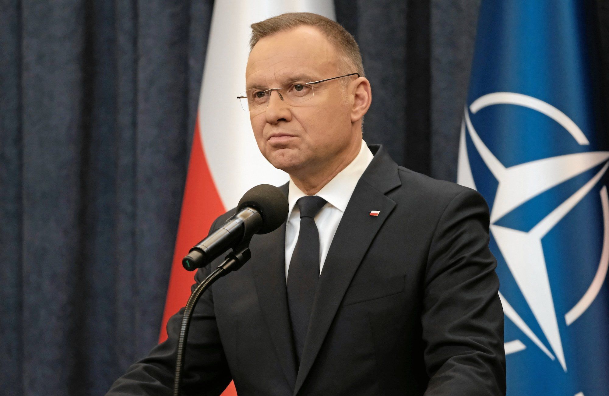Andrzej Duda w okularach stoi przy mikrofonie na tle flag Polski i NATO