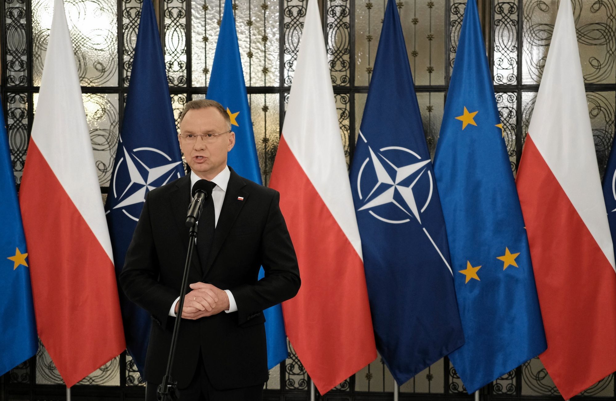 Andrzej Duda przemawia na tle flag Polski, Unii Europejskiej i NATO