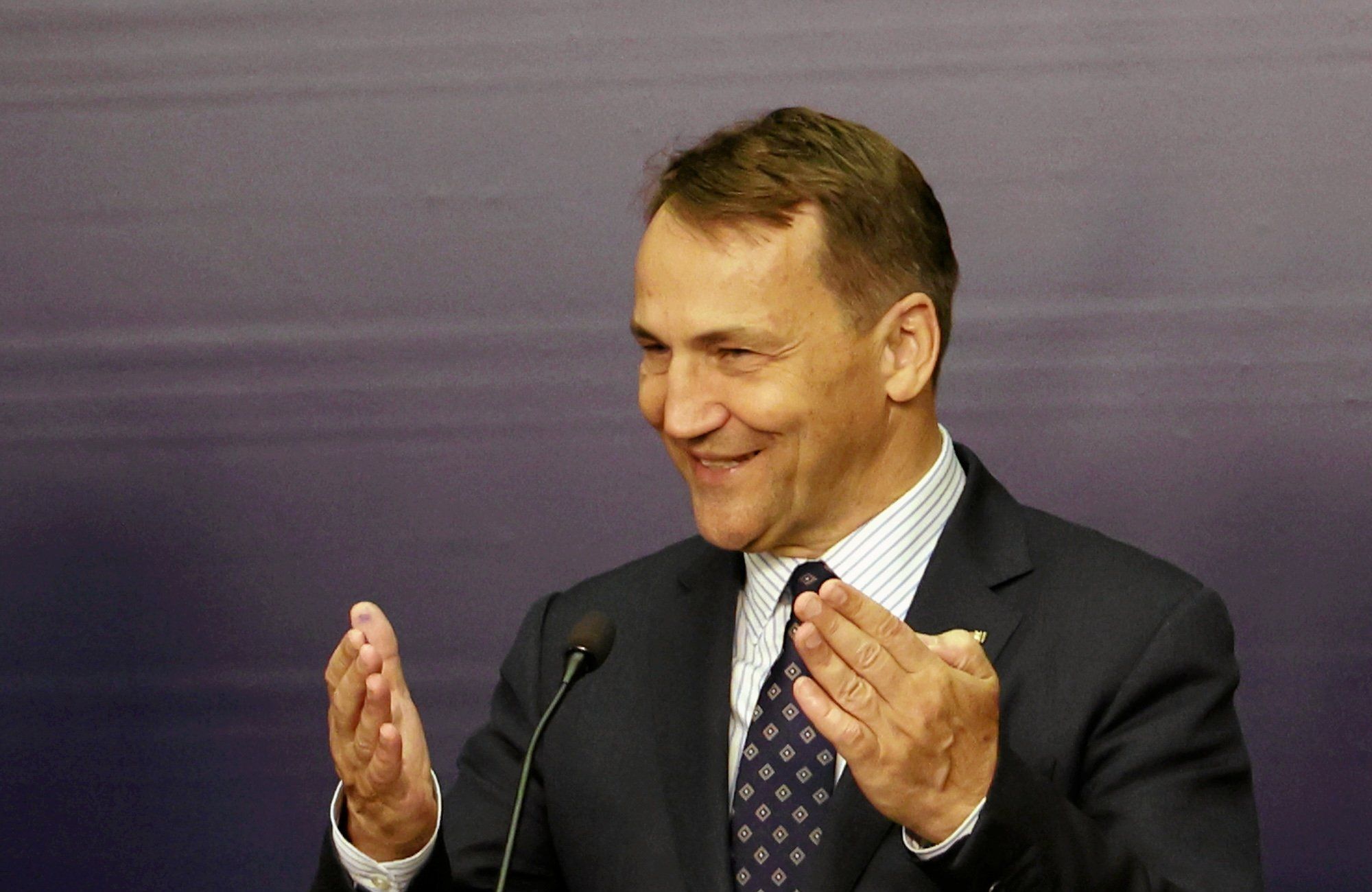 Radosław Sikorski przemawia do mikrofonu, unosząc dłonie. Uśmiecha się.