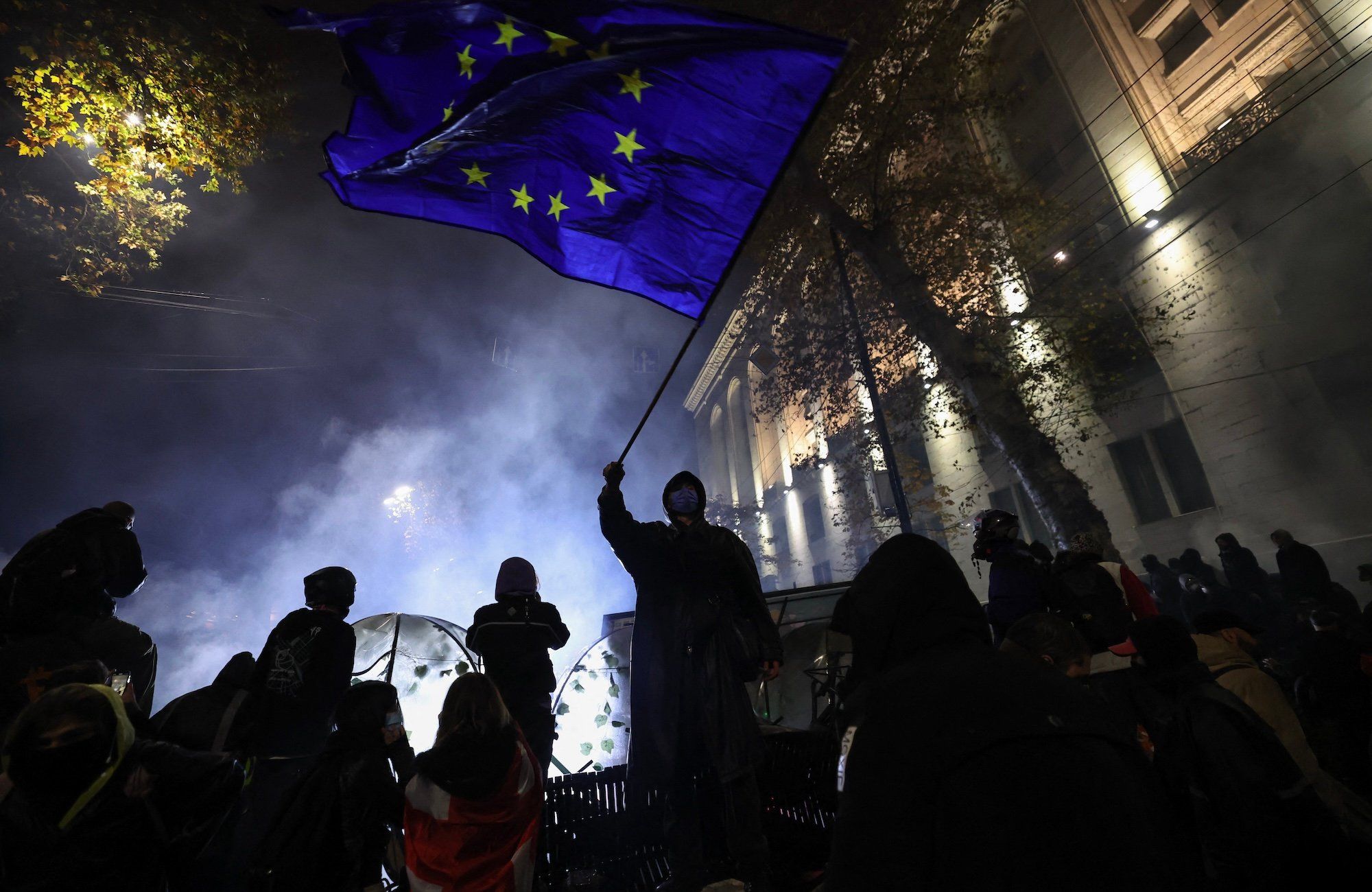 Uczestnicy nocnych protestów w Gruzji. Jedna z osób wymachuje dużą flagą Unii Europejskiej. W tle dym.