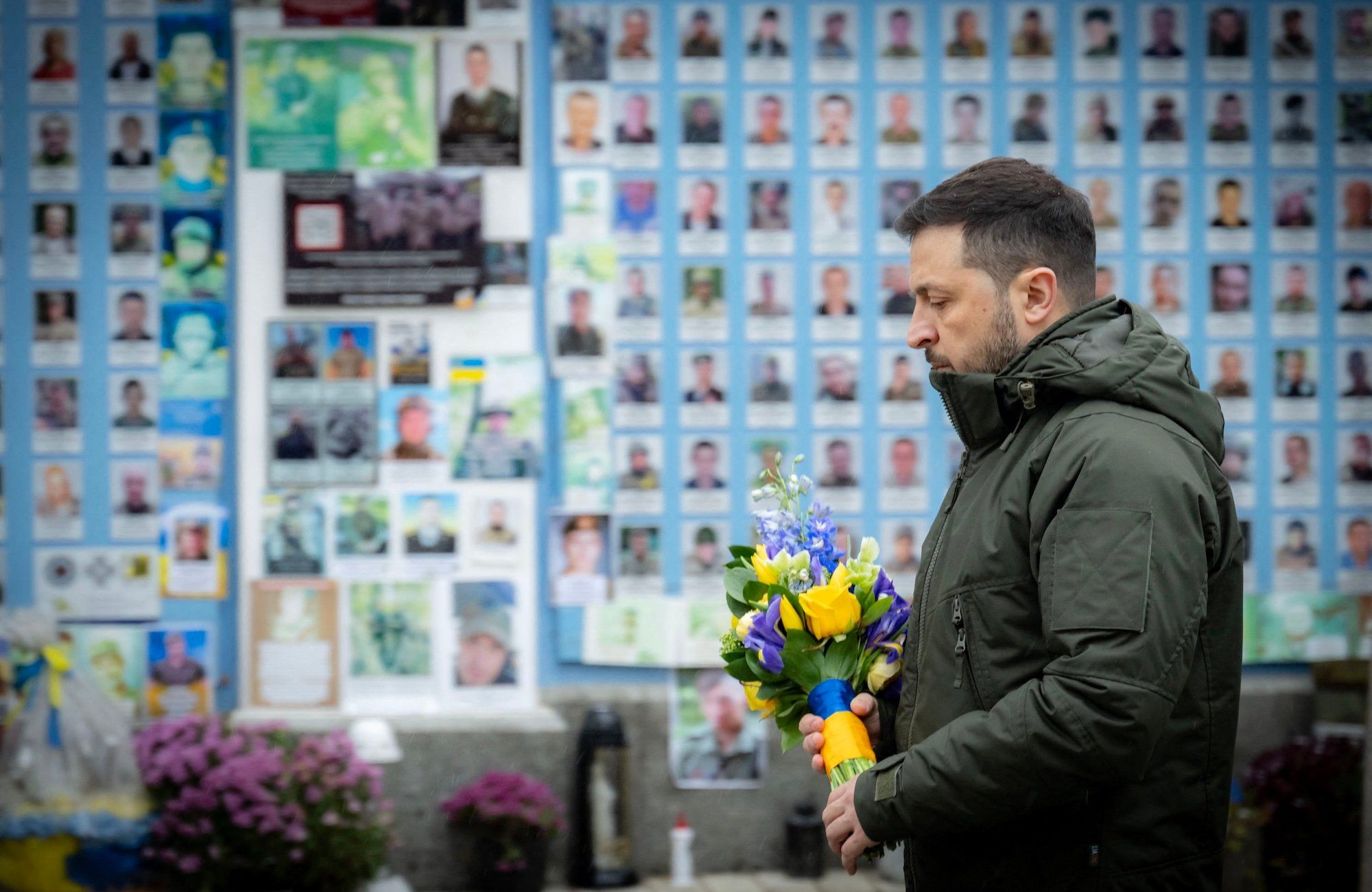 Wołodymyr Zełenski sfotografowany z lewego profilu, ubrany w zgniłozieloną kurtkę, w ręku trzyma bukiet żółtych i niebieskich kwiatów.