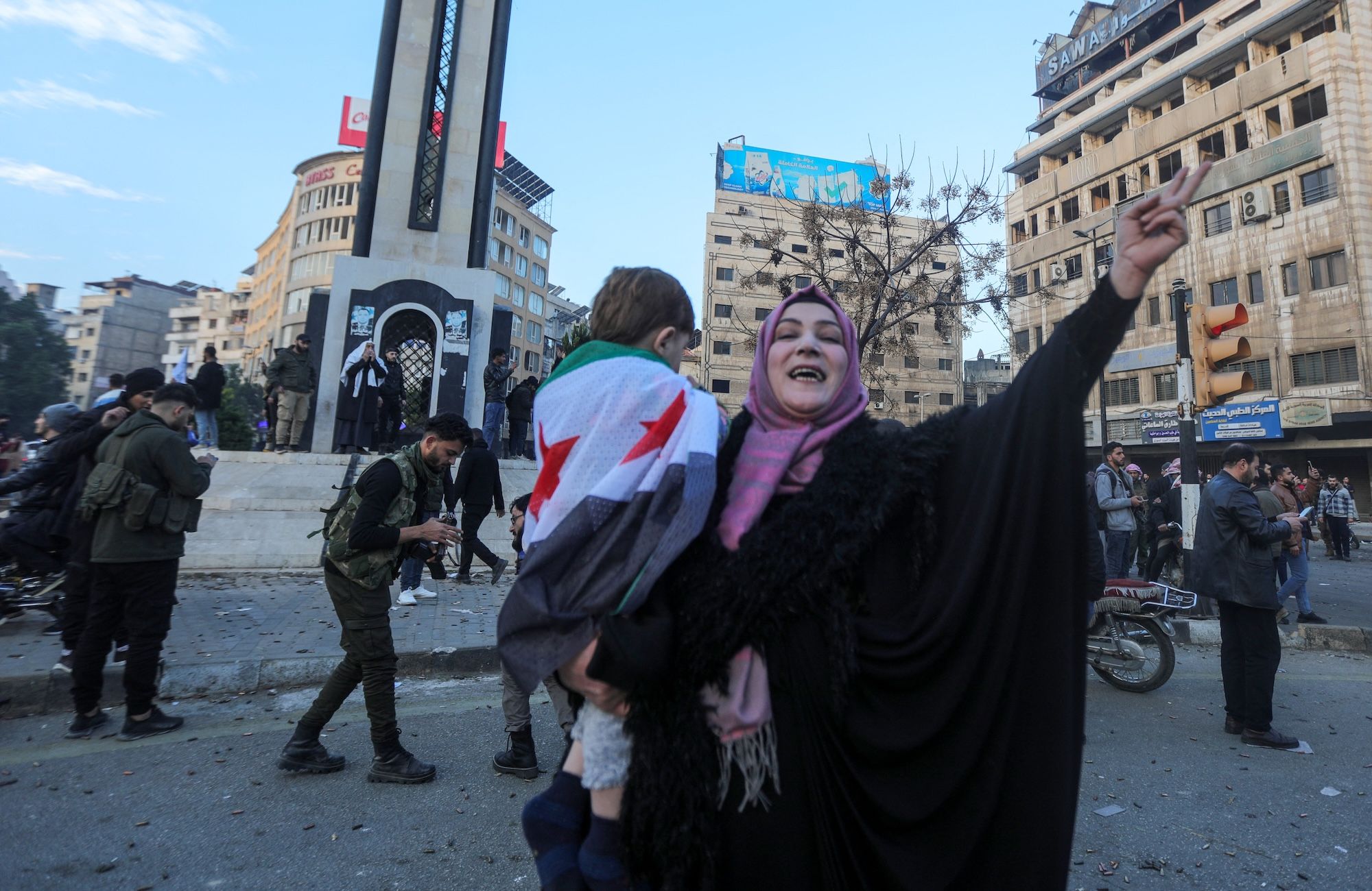 Syryjka w ciemnym stroju i różowej chuście na głowie trzyma na ręku dziecko owinięte we flagę Syrii. Unosi do góry rękę w geście wiktorii.