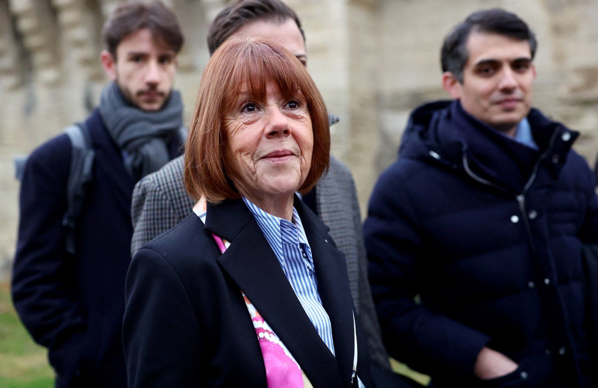 Gisele Pelicot w otoczeniu prawników. To 60-letnia kobieta w rudych włosach obciętych na boba, ubrana w niebieską koszulę i czarny płaszcz