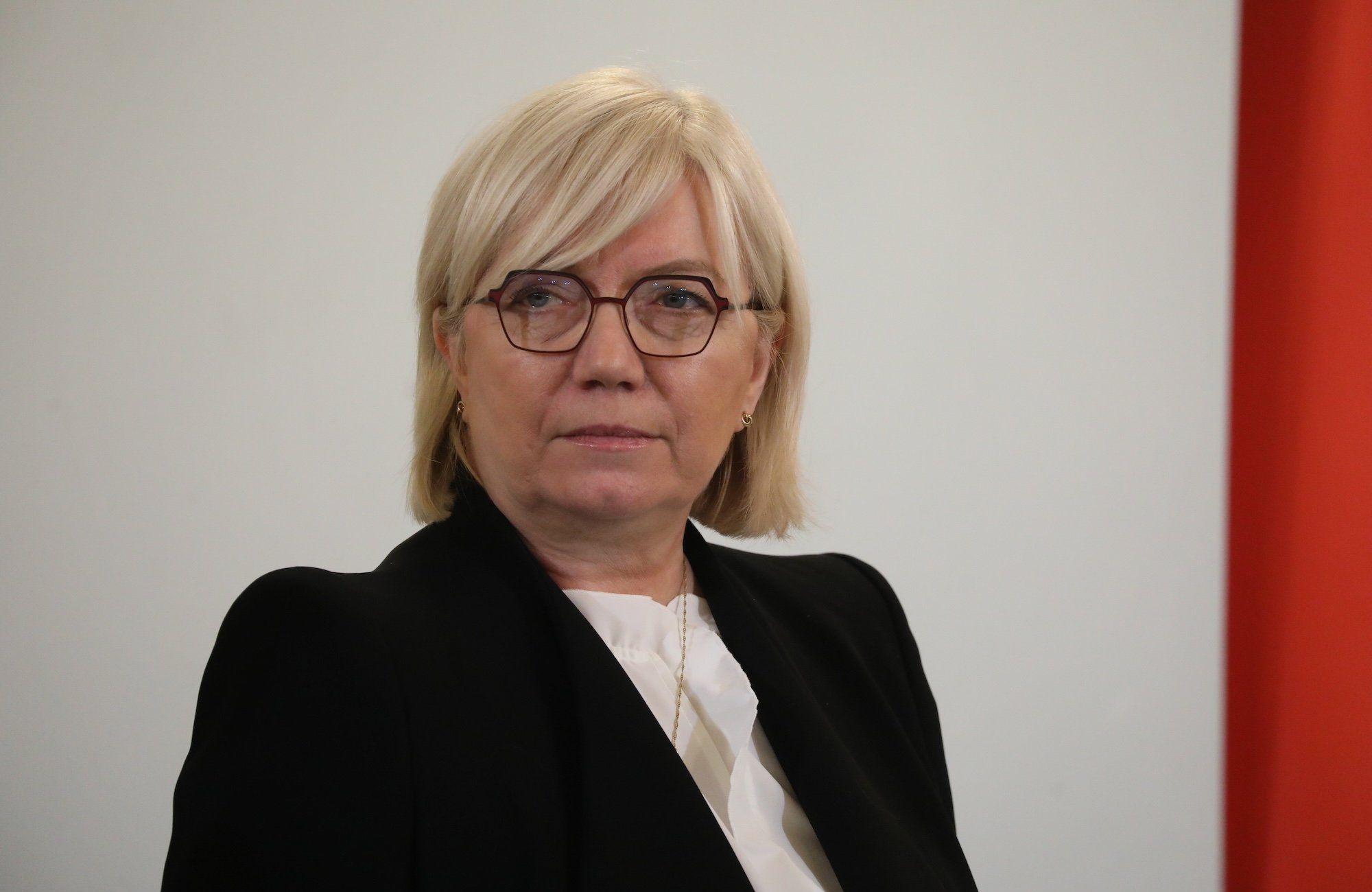 Portret Julii Przyłębskiej, ubranej w białą bluzkę i czarną marynarkę. Ma okulary w ciemnych oprawkach i jasne włosy
