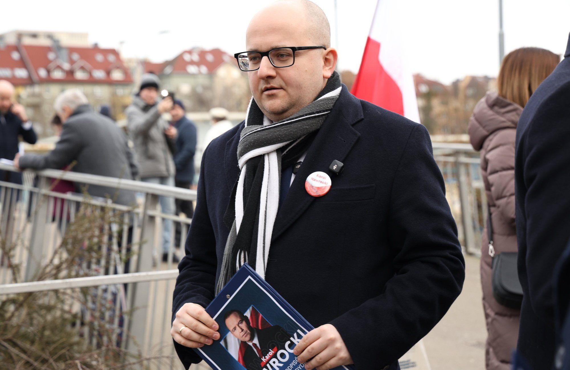 Poseł PiS Dariusz Matecki w ciemnym płaszczu i szaliku. Jest łysy i ma na twarzy okulary w ciemnych oprawkach. W ręku trzyma folder wyborczy Karola Nawrockiego
