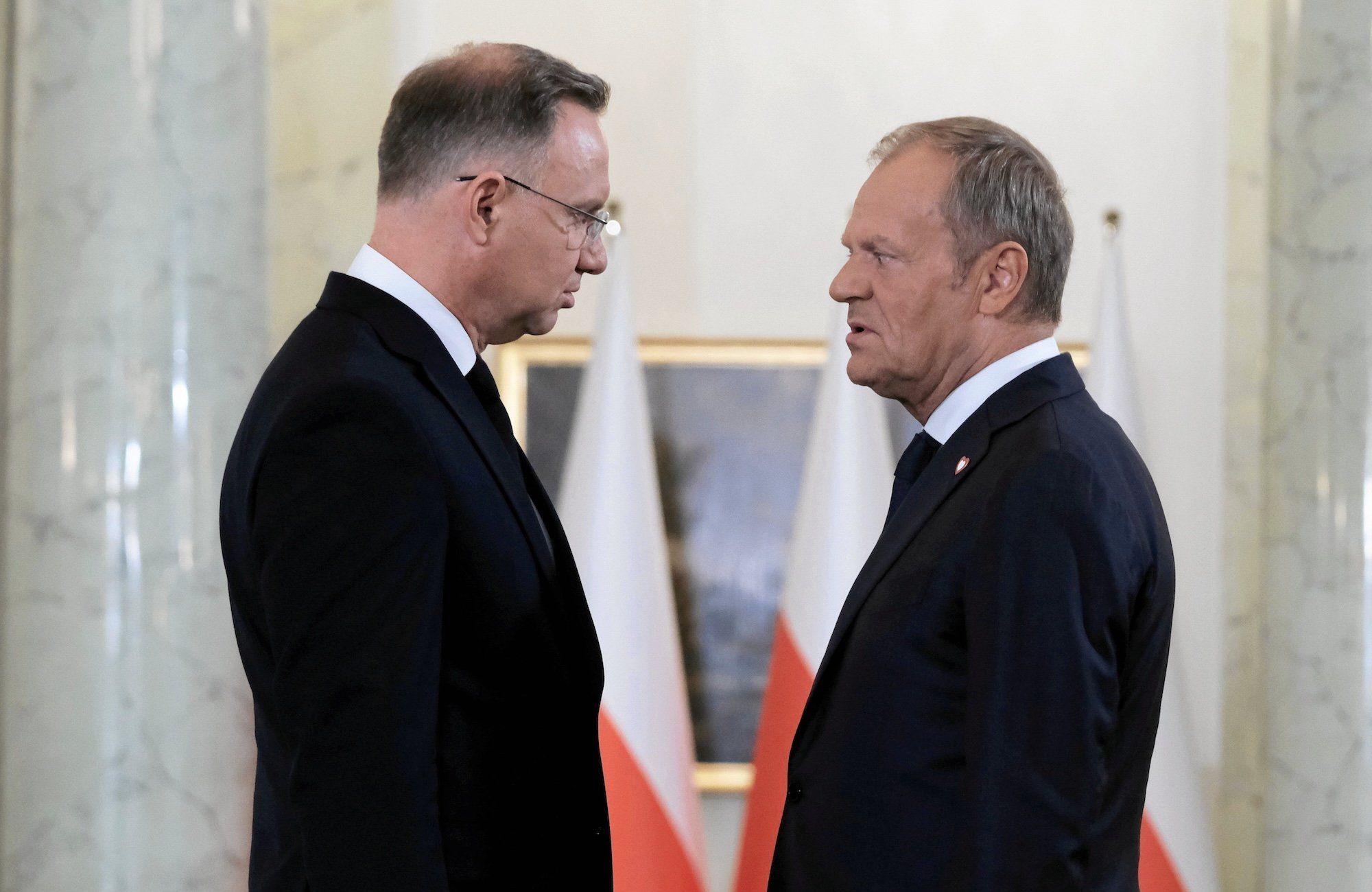 Andrzej Duda rozmawia z Donaldem Tuskiem w Pałacu Prezydenckim. Widziani z profilu na tle biało-czerwonych flag, patrzą na siebie wrogo