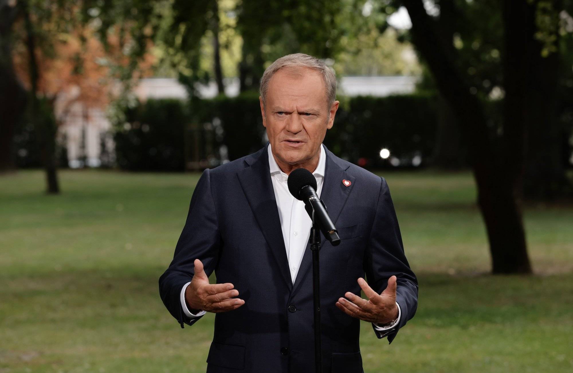 Donald Tusk przemawiający na konferencji prasowej. Ubrany w ciemną marynarkę i białą koszulę, bez krawata. W tle drzewa