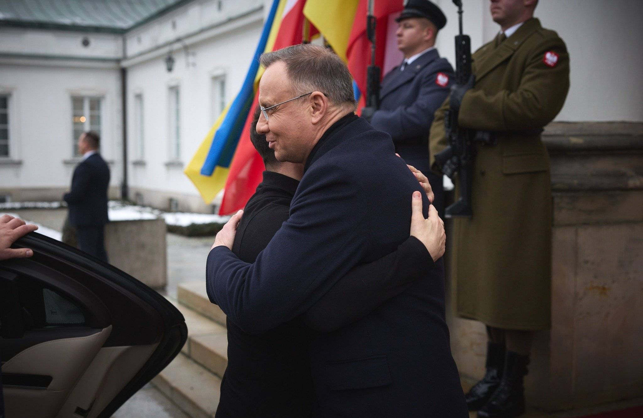 na zdjęciu jest czterech mężczyzn. Mężczyzna w czarnym płaszczu ściska mężczyznę w czarnej bluzie. Z tyłu stoi dwóch żołnierzy w mundurach polskiej armii