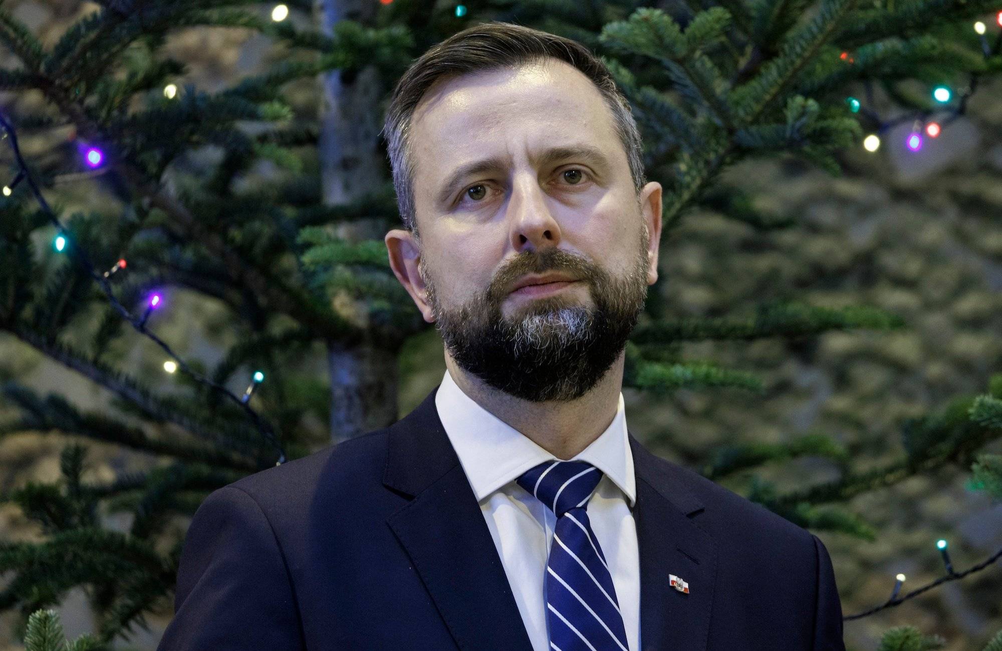 Władysław Kosiniak-Kamysz w granatowym garniturze na tle iglastego drzewa. Spogląda przed siebie z niepewną miną.