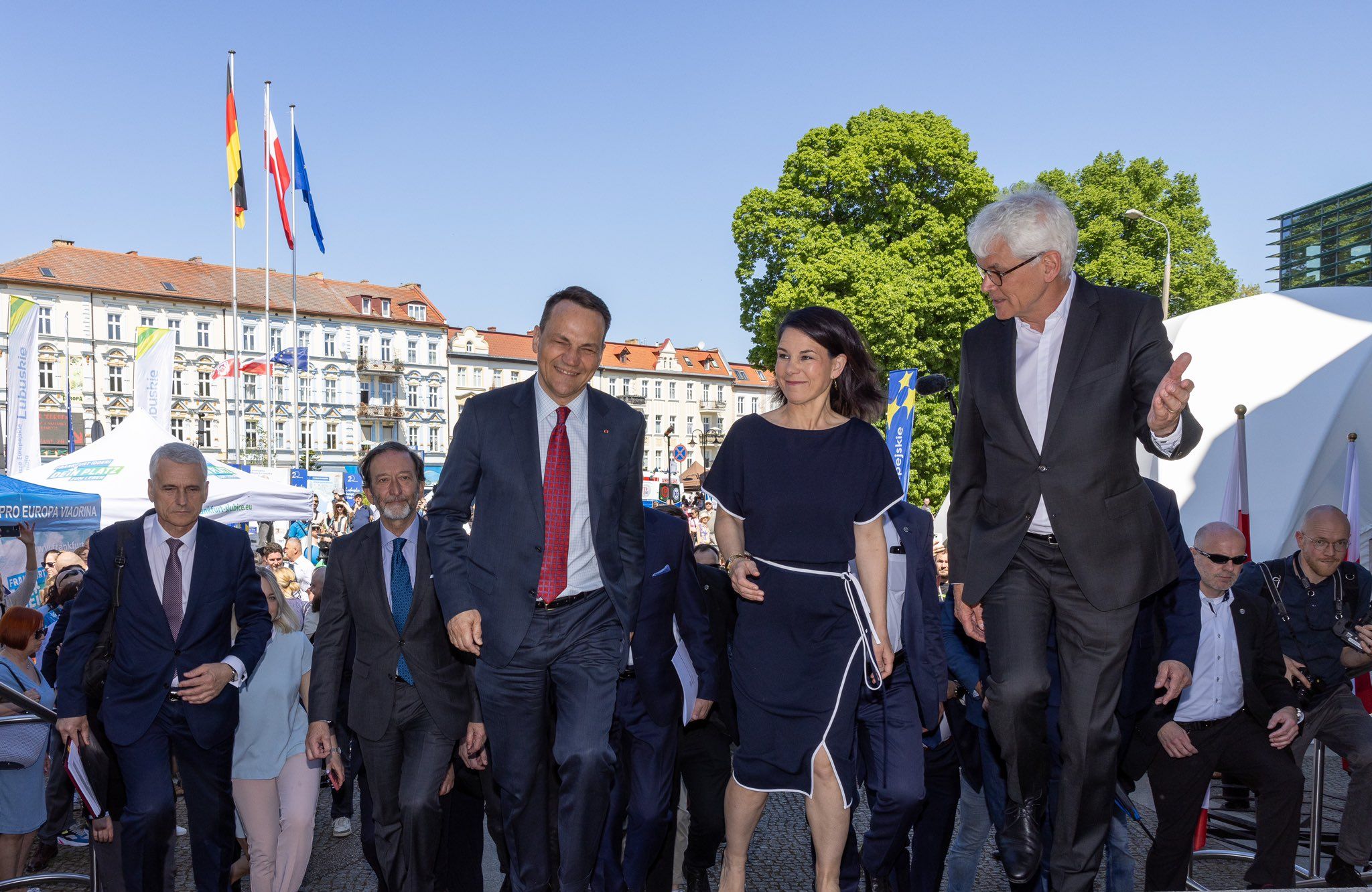Mężczyzna w garniturze i kobieta w eleganckiej, czarnej, prostej sukience z uśmiechami na twarzach zmierzają w stronę budynku, w którym zaraz odbędzie się konferencja prasowa. Towarzyszą im współpracownicy i organizatorzy oficjalnej wizyty. Zdjęcie zrobione na zewnątrz, jest ładna pogoda.
