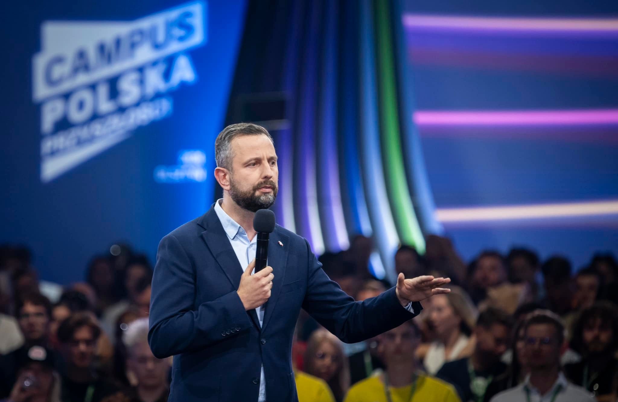 Brodaty mężczyzna (Władysław Kosiniak-Kamysz) w granatowym garniturze i białej koszuli z rozpiętym kołnierzykiem przemawia do mikrofonu. Za nim biały napis na niebieskim tle "Campus Polska".