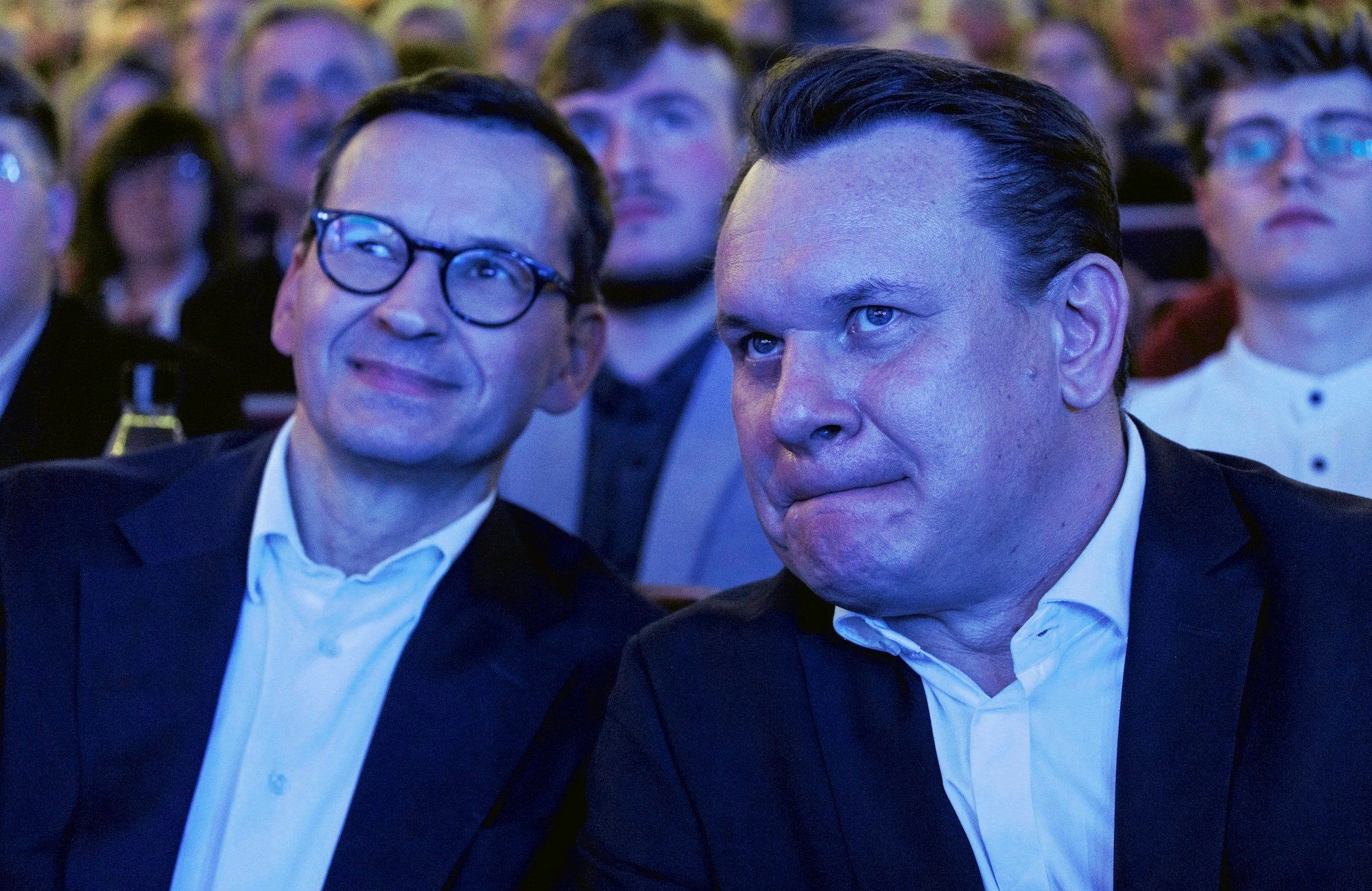 Mateusz Morawiecki i Dominik Tarczyński słuchają konferencji, spowici niebieską poświatą ekranu