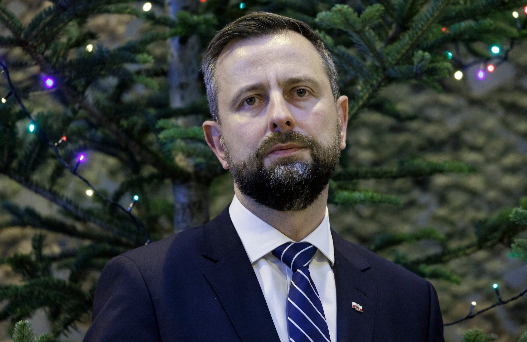 Władysław Kosiniak-Kamysz w granatowym garniturze na tle iglastego drzewa. Spogląda przed siebie z niepewną miną.
