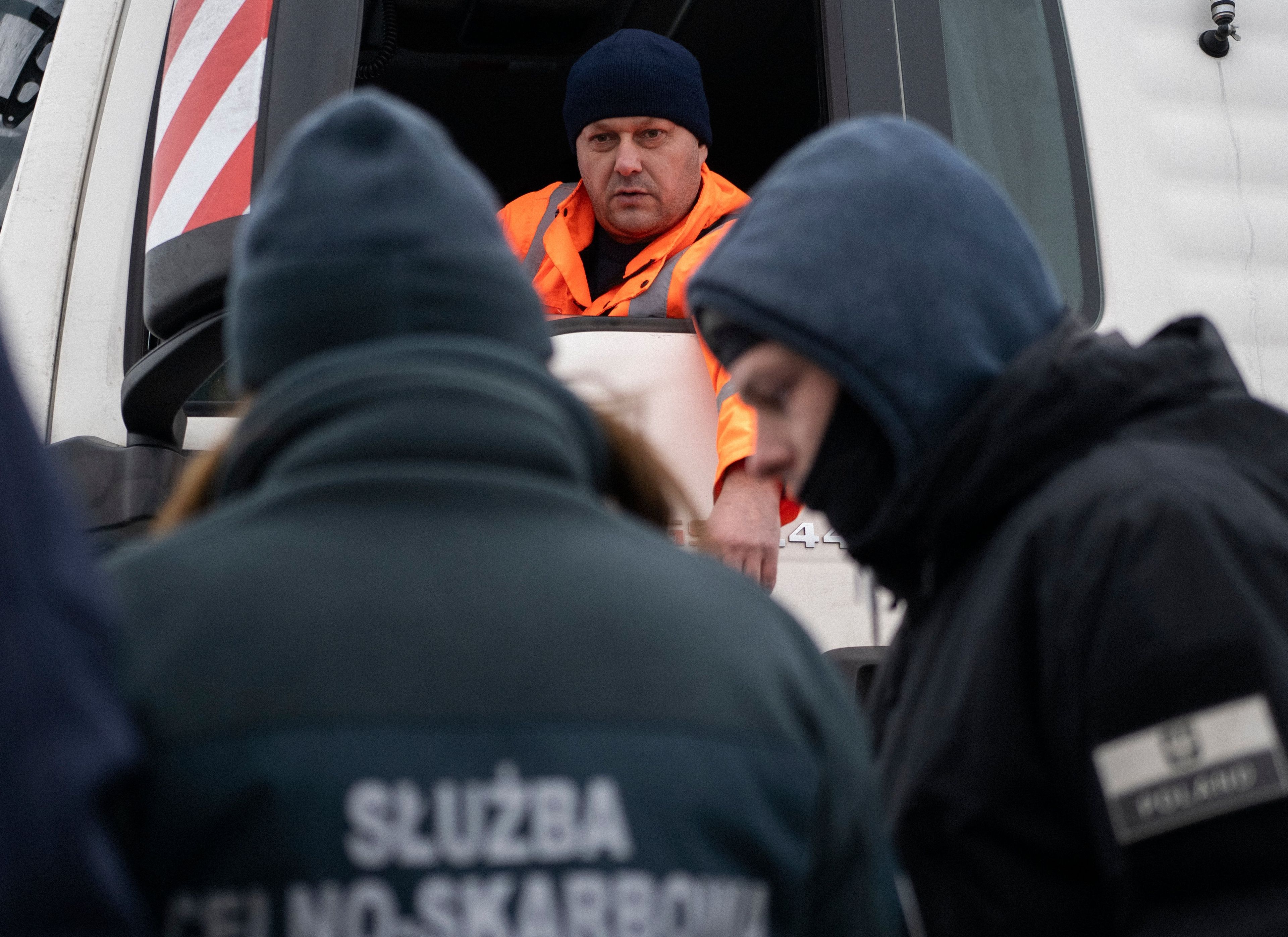 Kontrola graniczna, na pierwszym planie funkcjonariusze służby celnej pochyleni nad dokumentami, w głębi kierowca w kabinie ciężarówki. Blokada na granicy