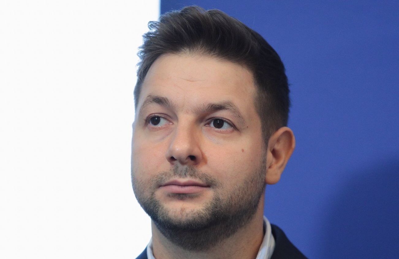Patryk Jaki, europoseł Solidarnej Polski spogląda w prawo spod uniesionych brwi. Ubrany w ciemną marynarkę i rozpiętą pod szyją koszulę.