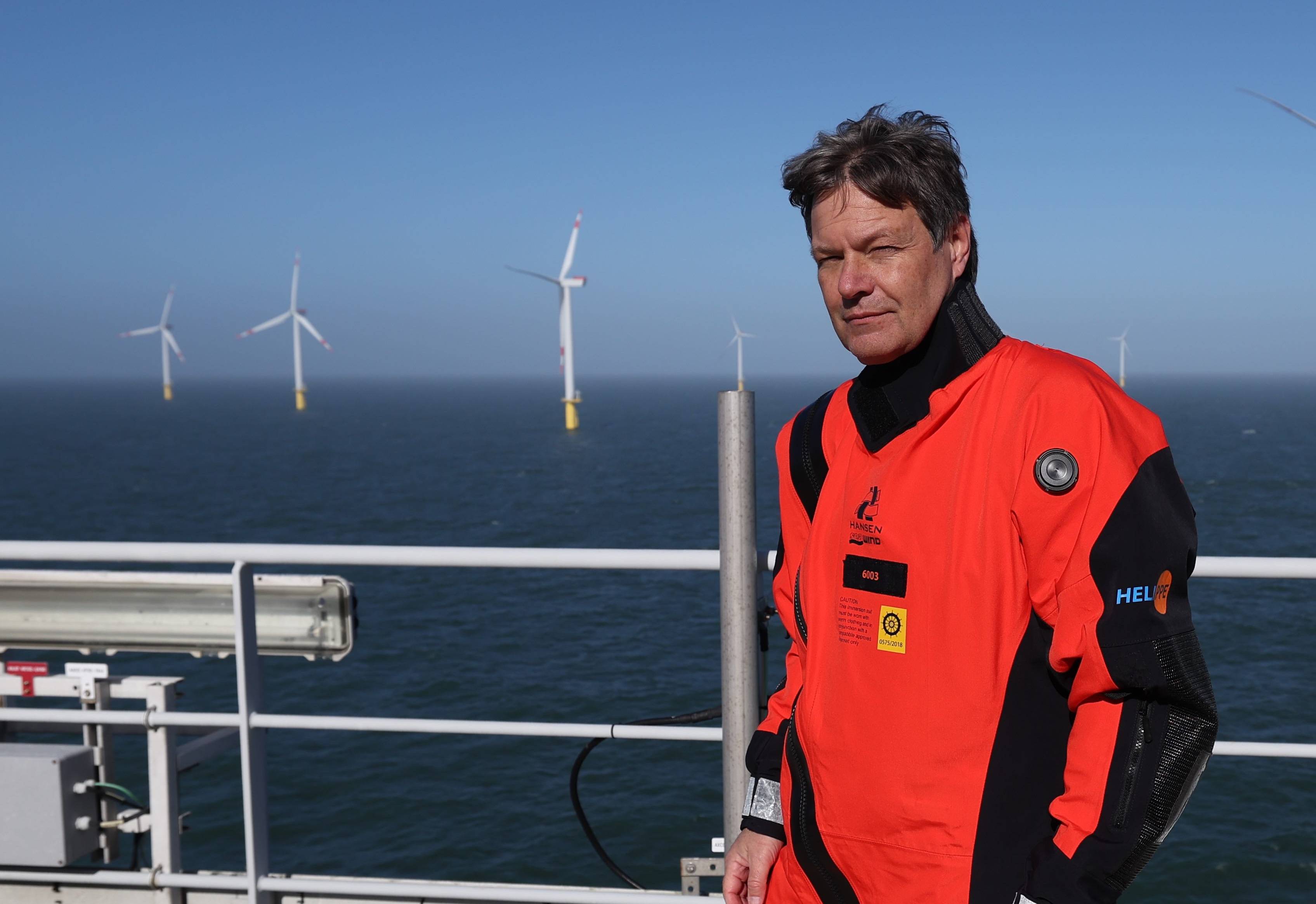 Robert Habeck w czerwonym kombinezonie stoi na tle elektrowni wiatrowych, zbudowanych w morzu.