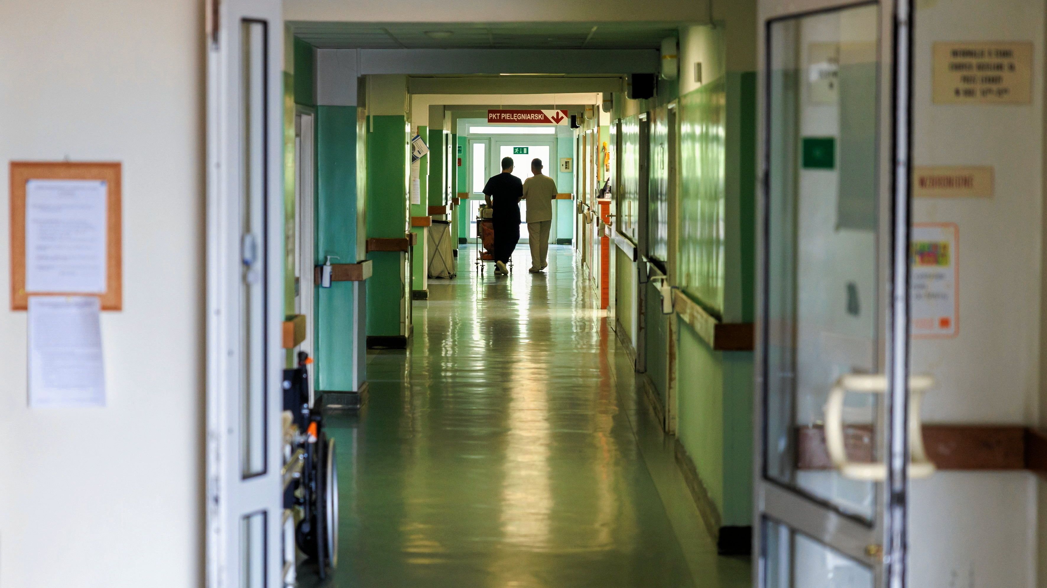 Na zdjęciu jest korytarz w szpitalu, w kolorach jasno zielonych, w końcu korytarza widać dwie osoby