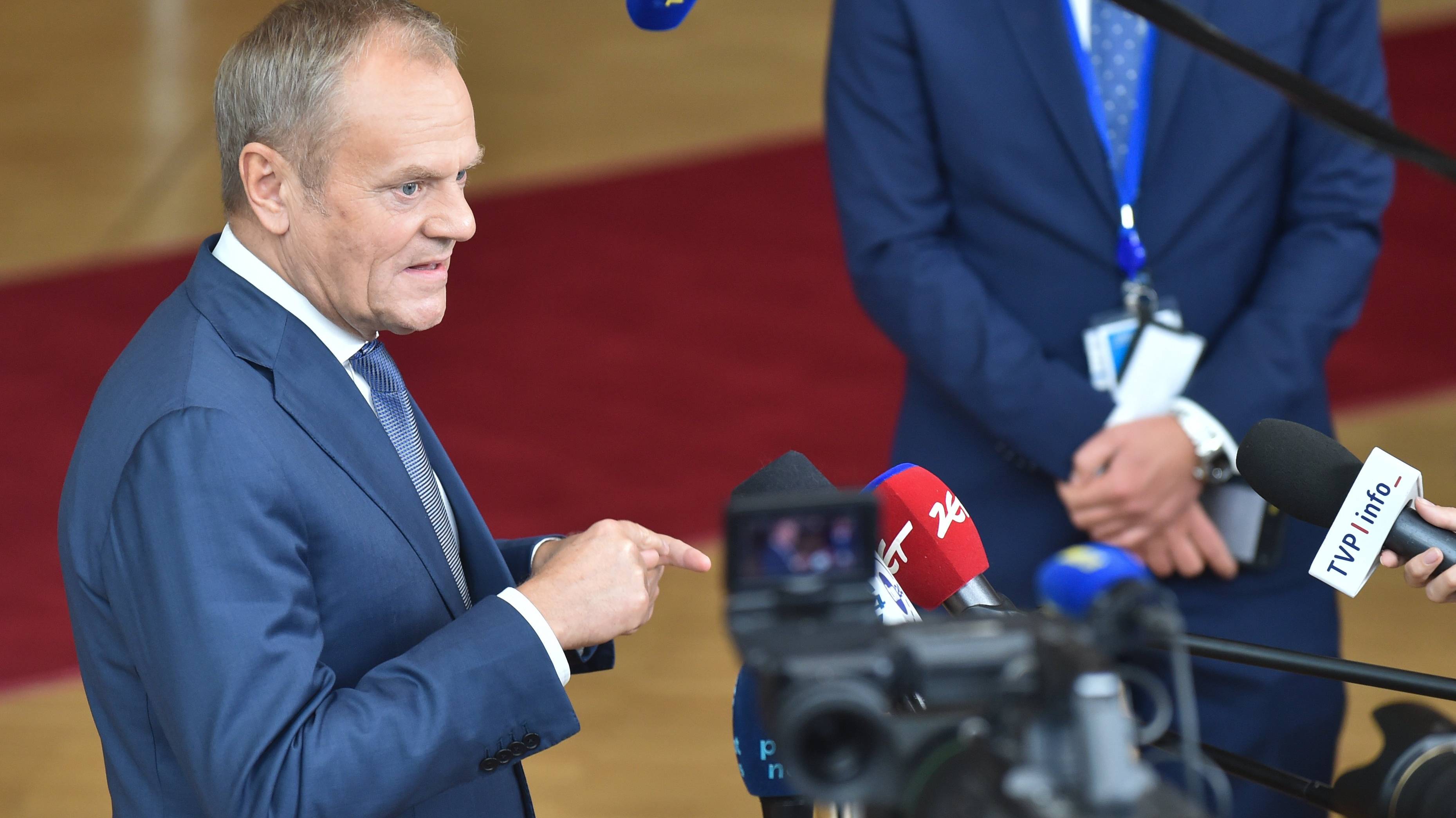 Premier Donald Tusk w niebieskim garniturze stoi przed mikrofonami dziennikarzy w holu prowadzącym do sali plenarnej budynku Rady Europejskiej, w tle widać czerwony dywan.