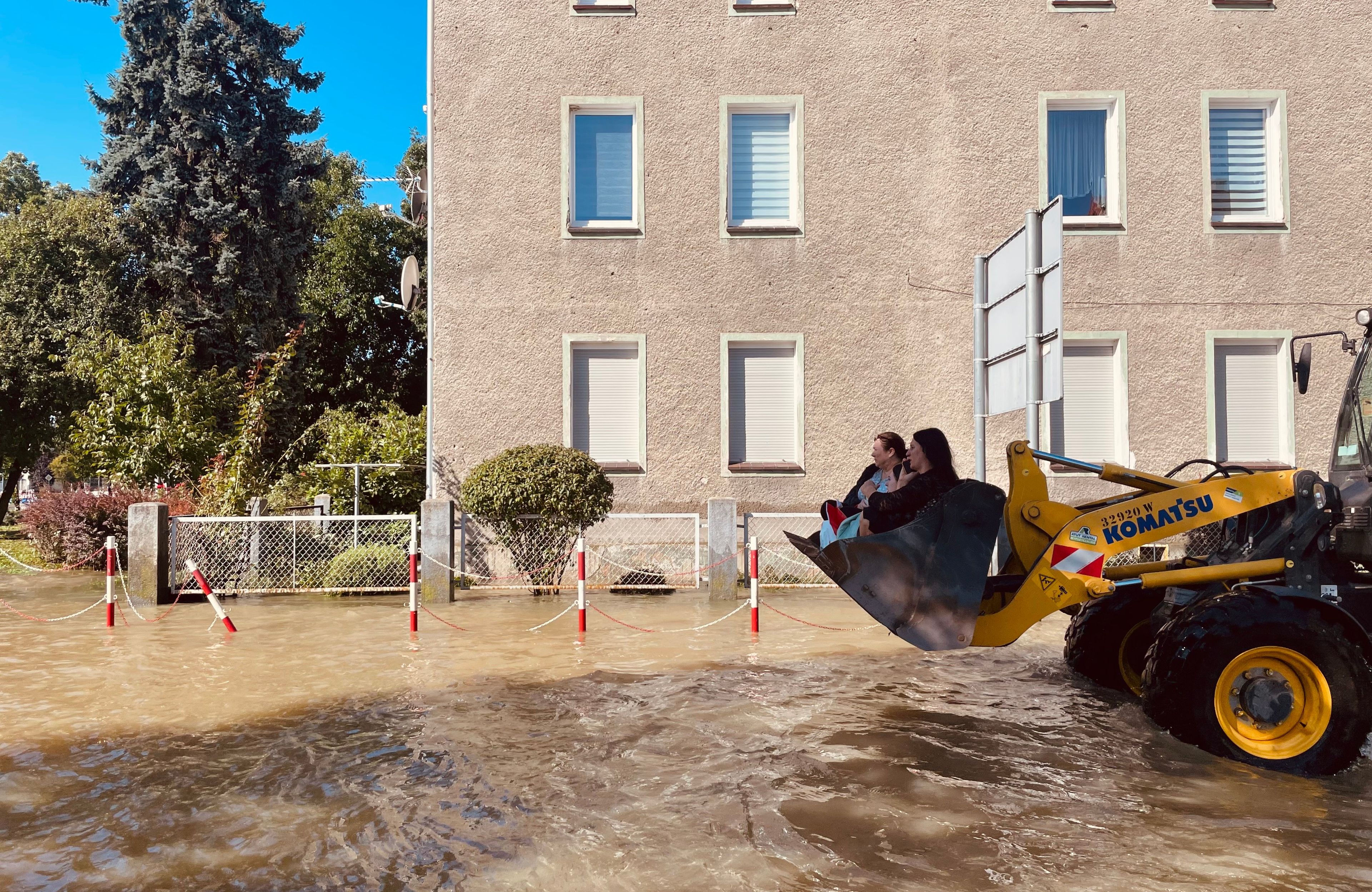 Kobiety siedzą w łyżce koparki, która jedzie zalaną wodą ulicą