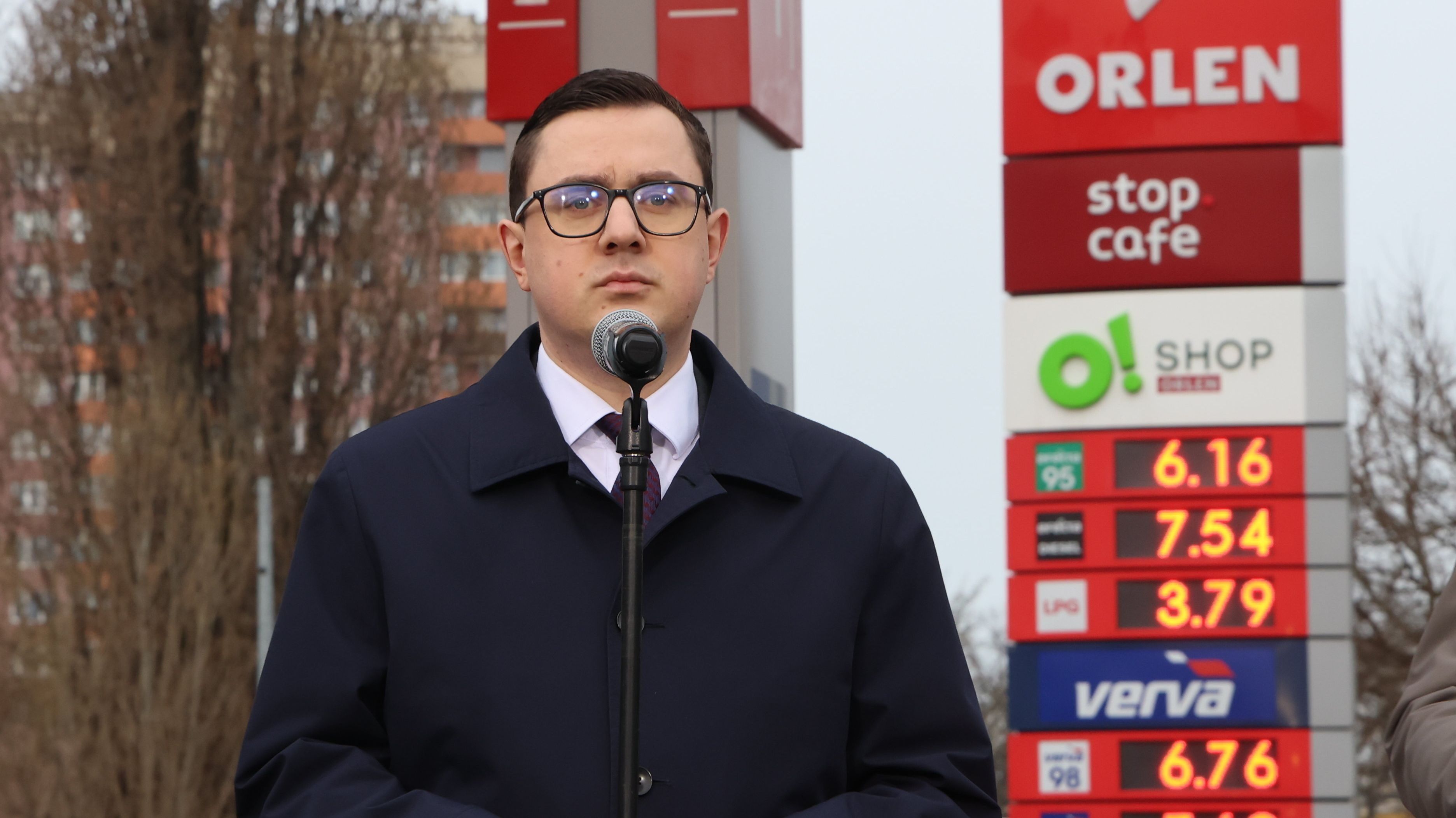 minister Miłosz Mityka w garniturze i płaszczu na tle stacji Orlen