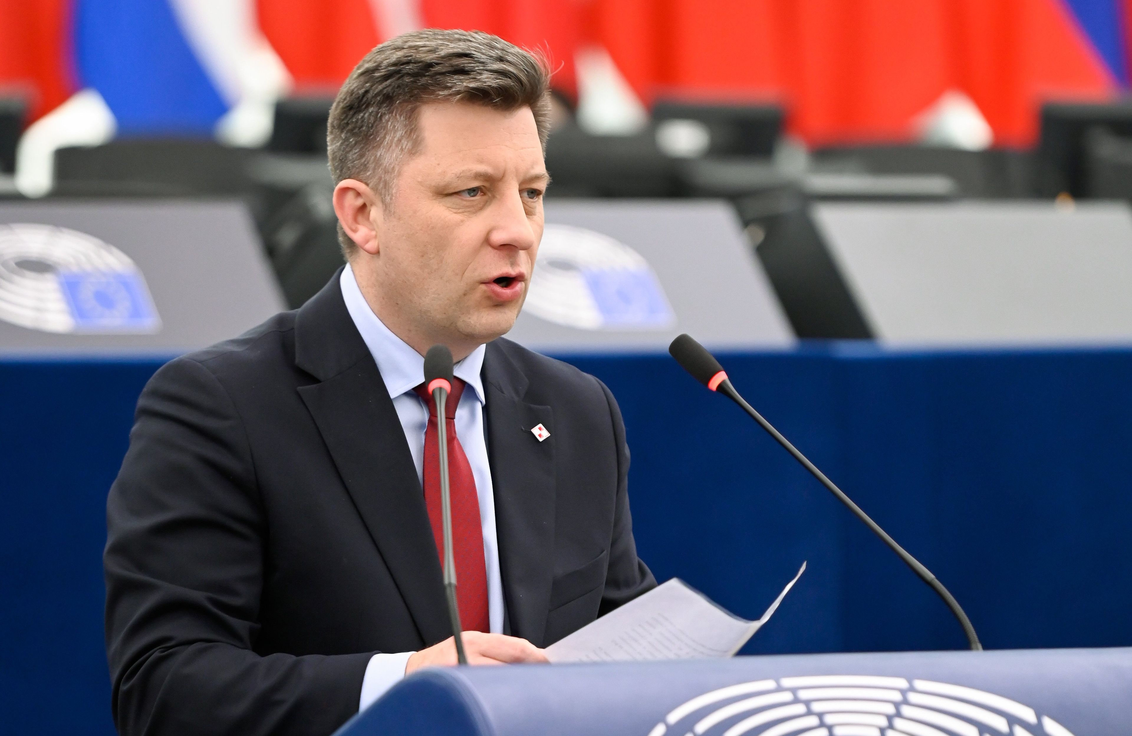 Mężczyzna w średnim wieku z ciemnoblond włosami, w ciemnogranatowym garniturze z czerwonym krawatem stoi za niebieską mównicą i mówi do mikrofonu, w tle zarys rzędu kolorowych flag.