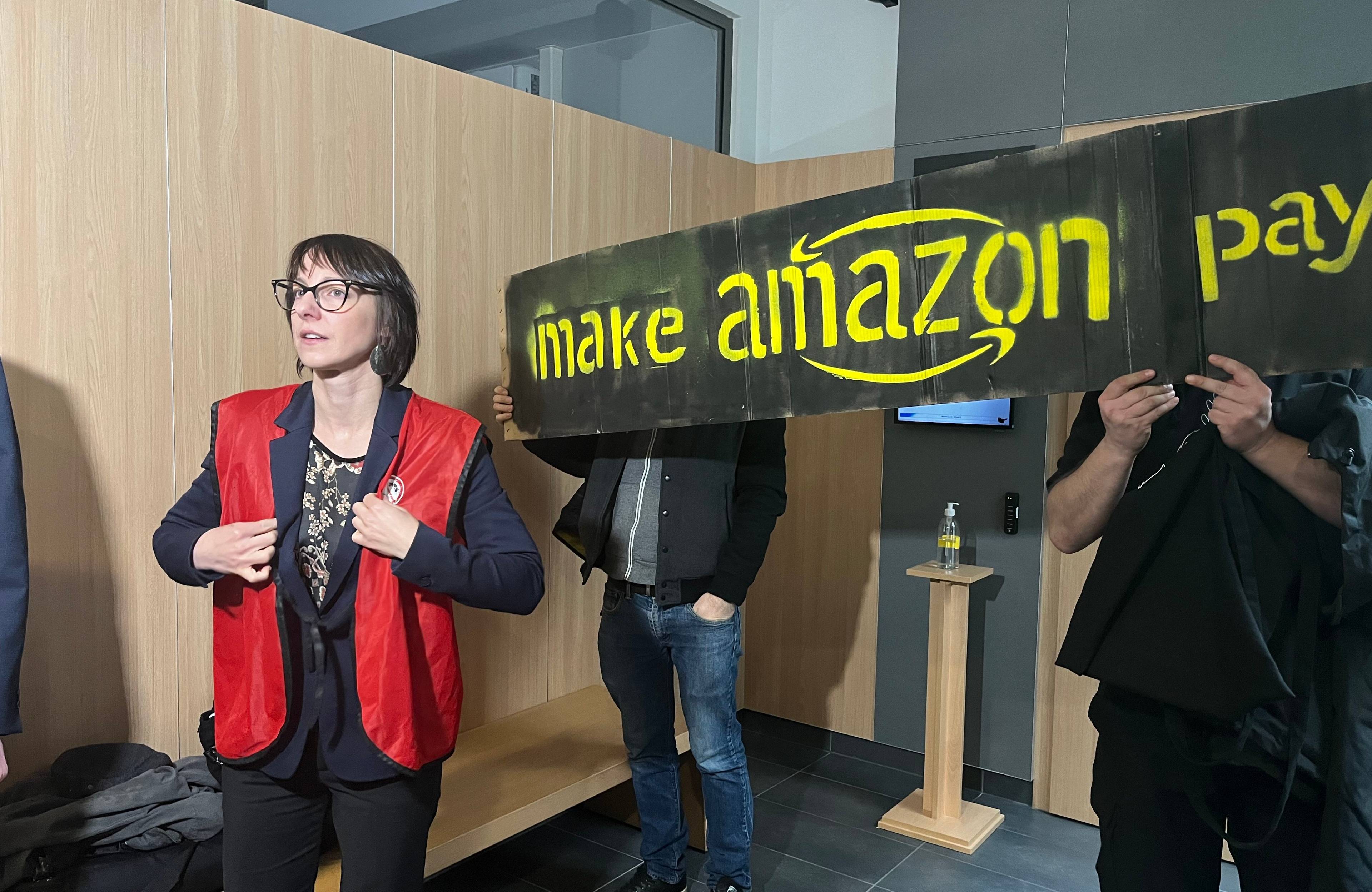Kobieta w czerwonej kamizelce na tle transparentu "Make Amazon pay"