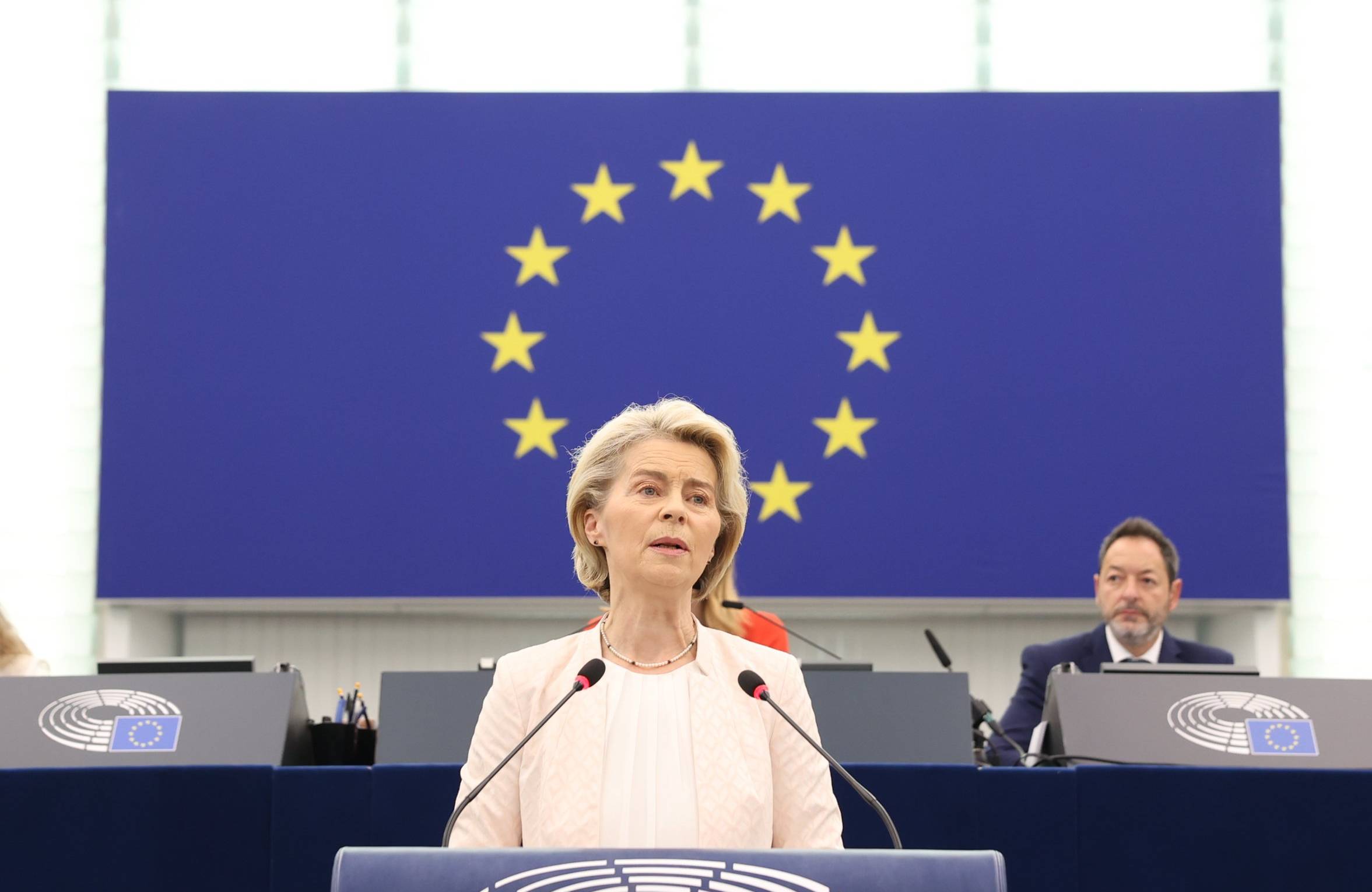 Kobieta z krótkimi blond włosami, jasnej, różowej marynarce, stoi przed mikrofonem, w tle niebieska flaga z żółtymi gwiazdkami