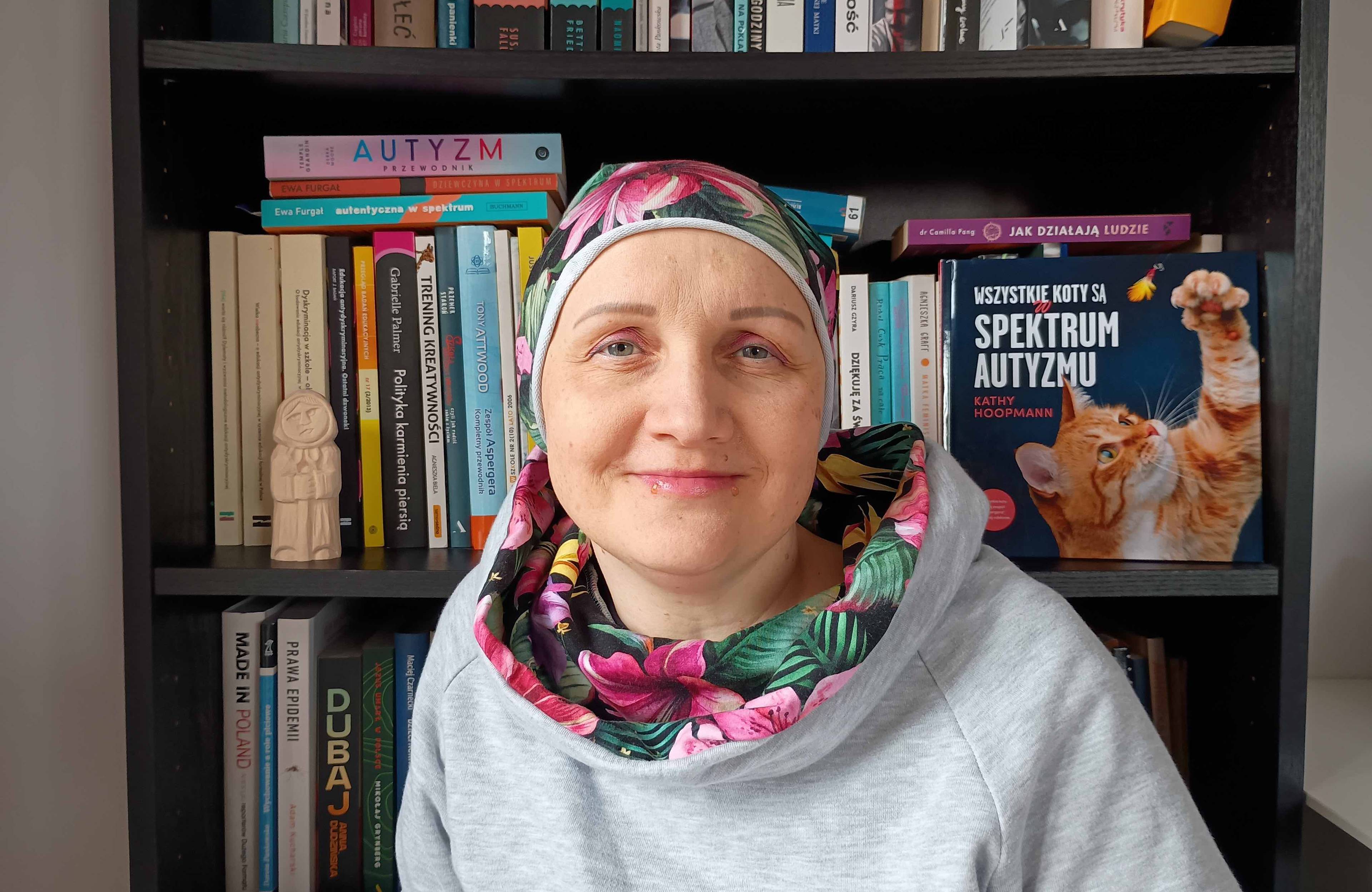 Kobieta w chustce na głowie na tle biblioteczki, na półce stoi książka pod tytulwem Wszystkie koty są w spektrum autyzmu