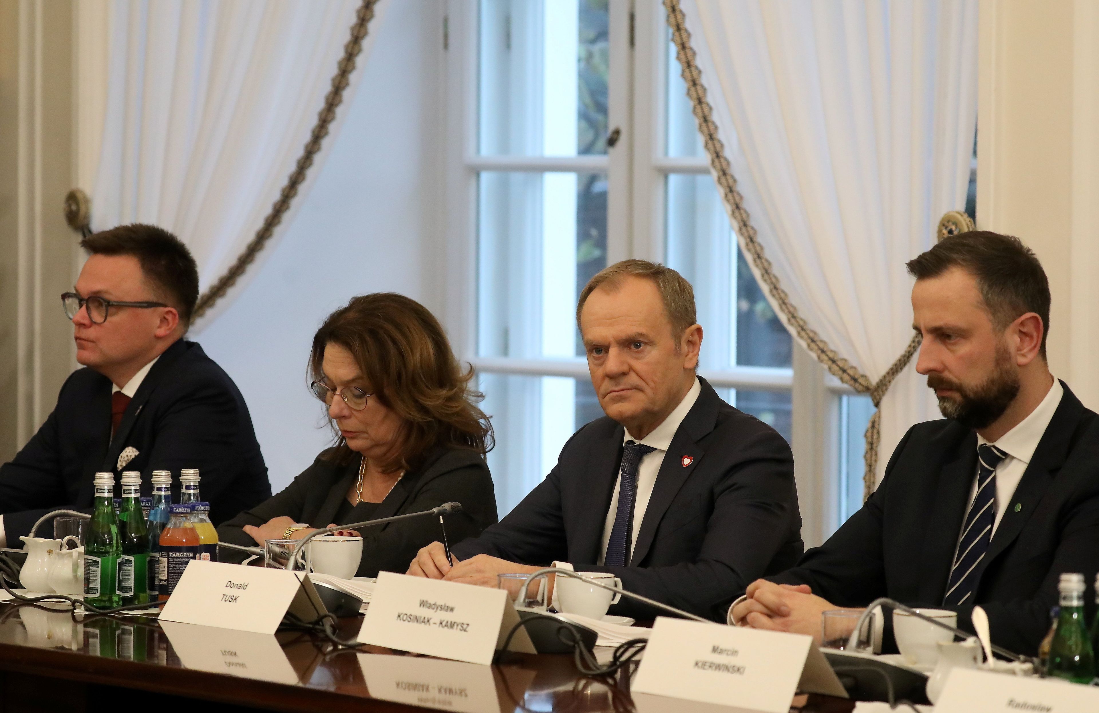 Politycy koalicji rządowej siedzą za długim stołem, przed nimi tabliczki z nazwiskami