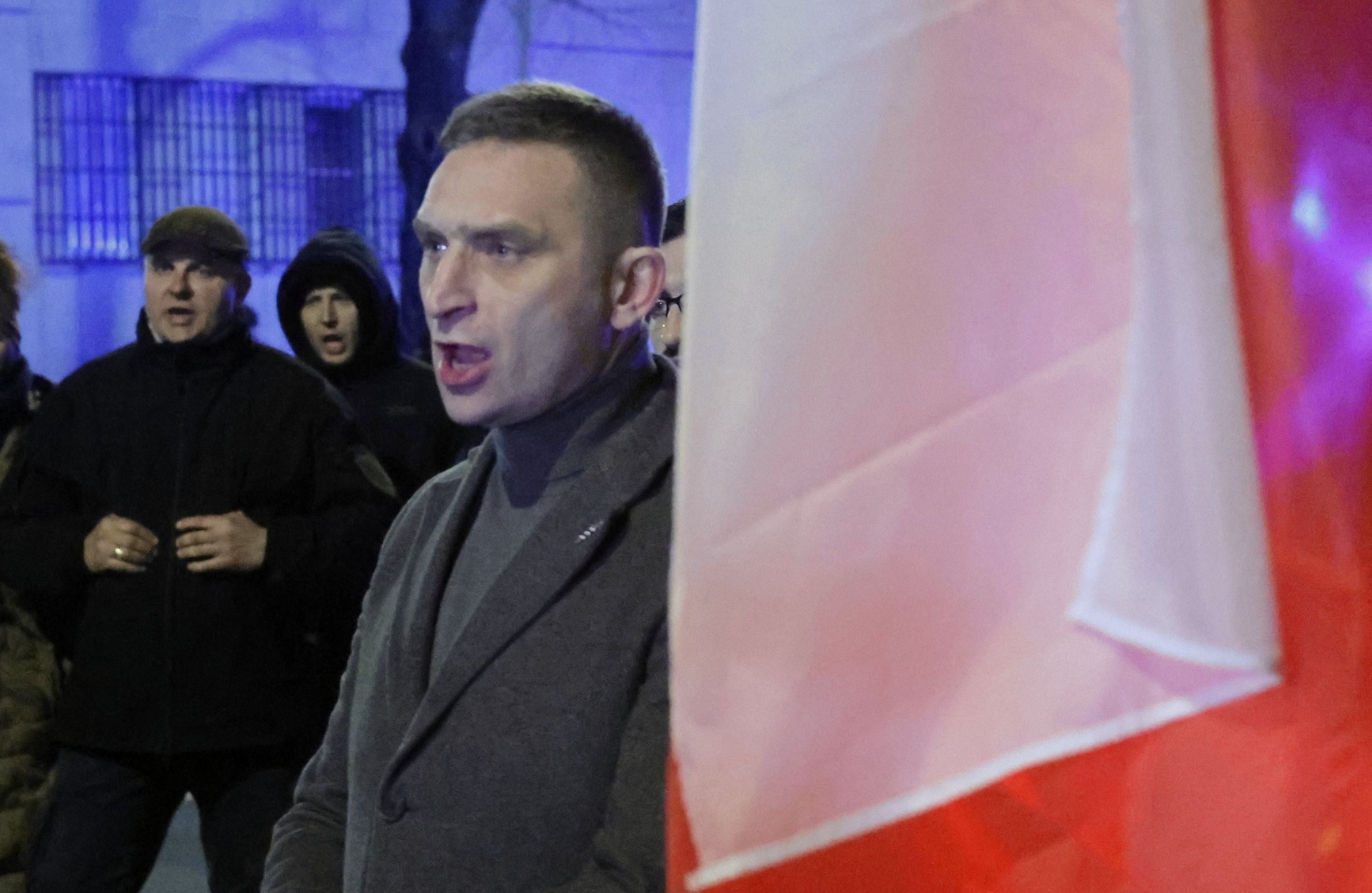 Ubrany w płaszcz mężczyzna krzyczy na ulicy, obok niego polska flaga